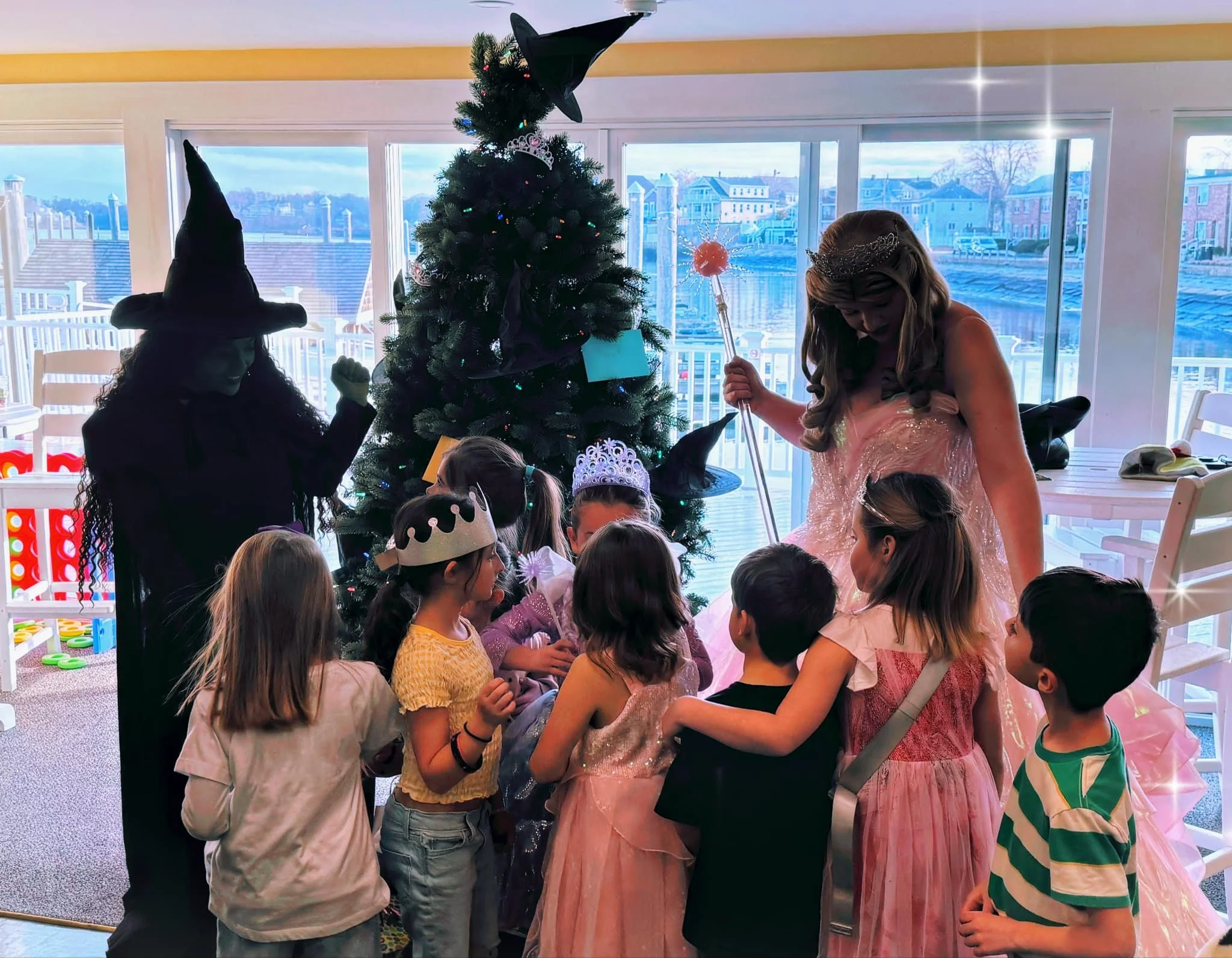 Children dressed as princesses and a witch gather around a Christmas tree with the Oz Witches Party Princesses during a birthday party in a bright room with large windows.
