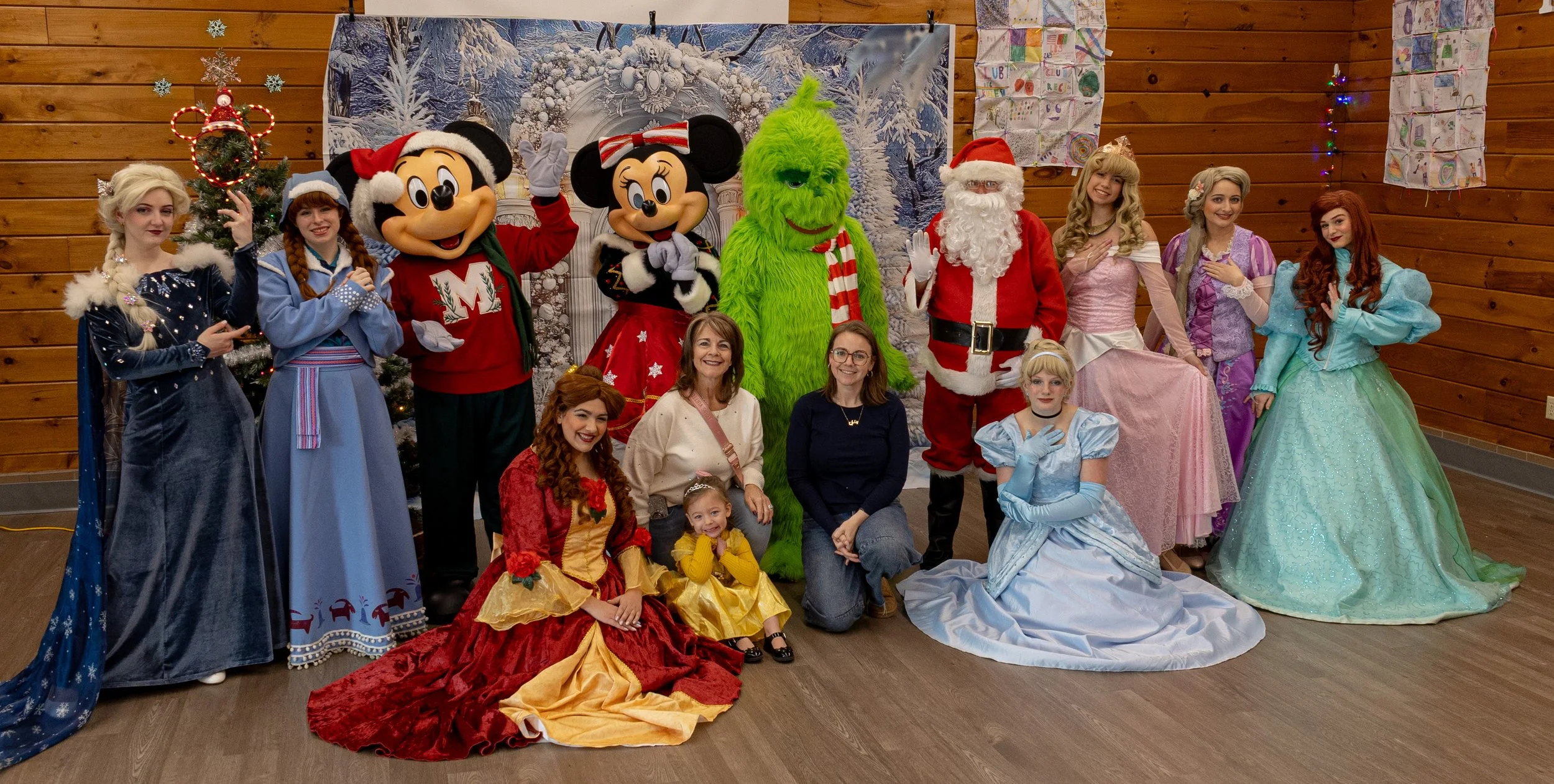Party Princesses and Characters posing with children in holiday costumes, inside a wooden room with holiday decorations and a snow-themed backdrop.