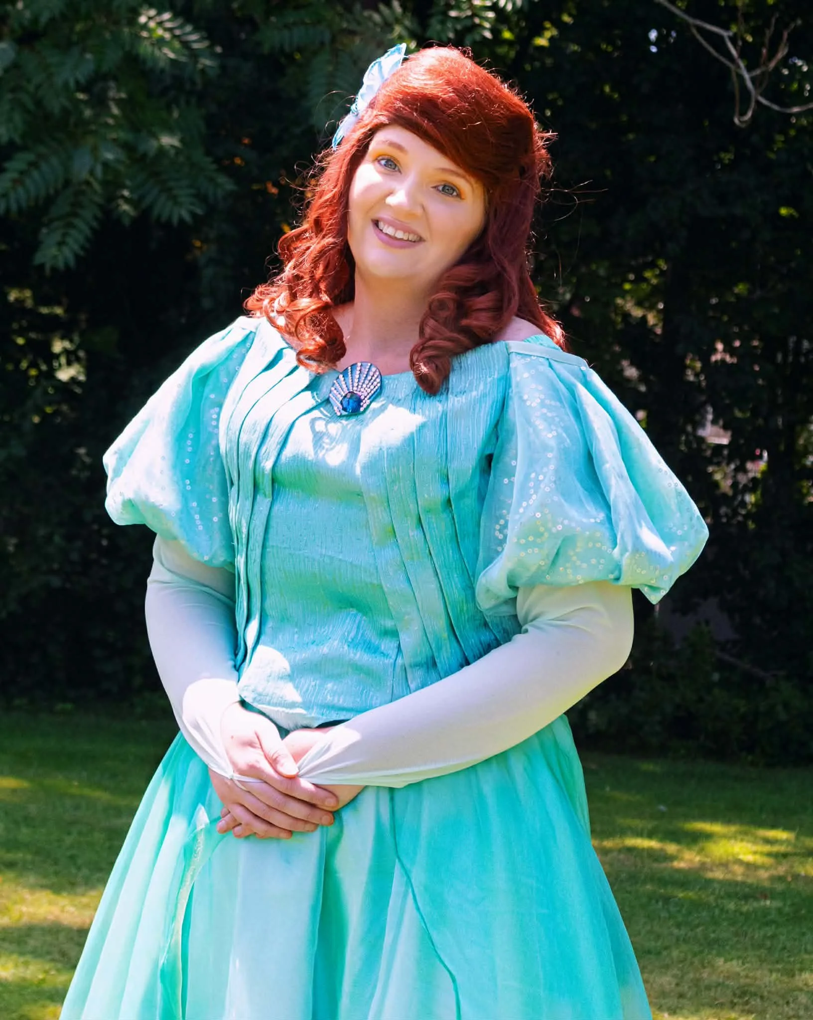 The Little Mermaid party princess standing on grass in front of trees, wearing a light blue gown with green puffed sleeves and long red hair.