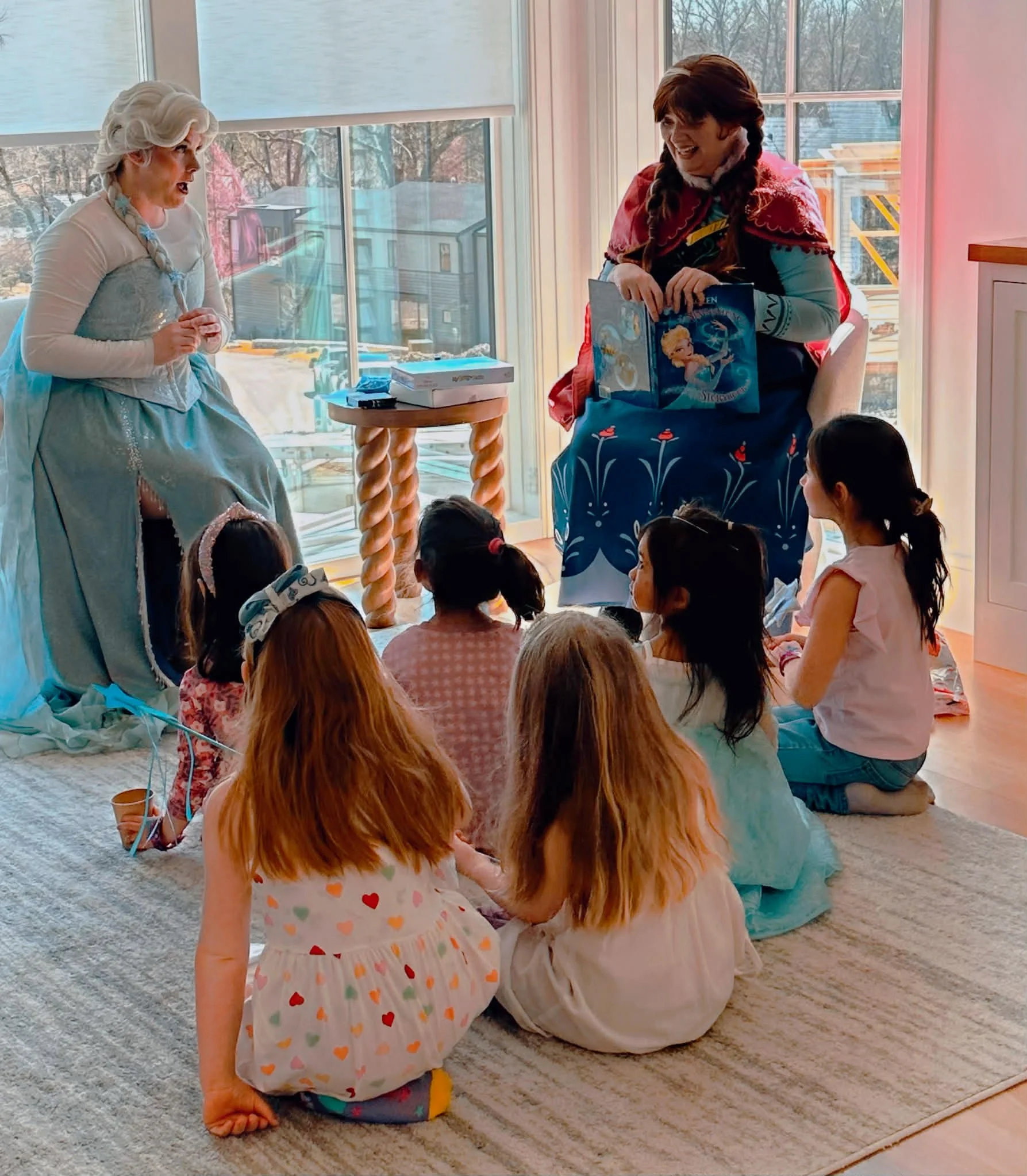 Children sitting on the carpet and listening to a woman in a princess costume tell a story, while another woman in a cape holds a book.