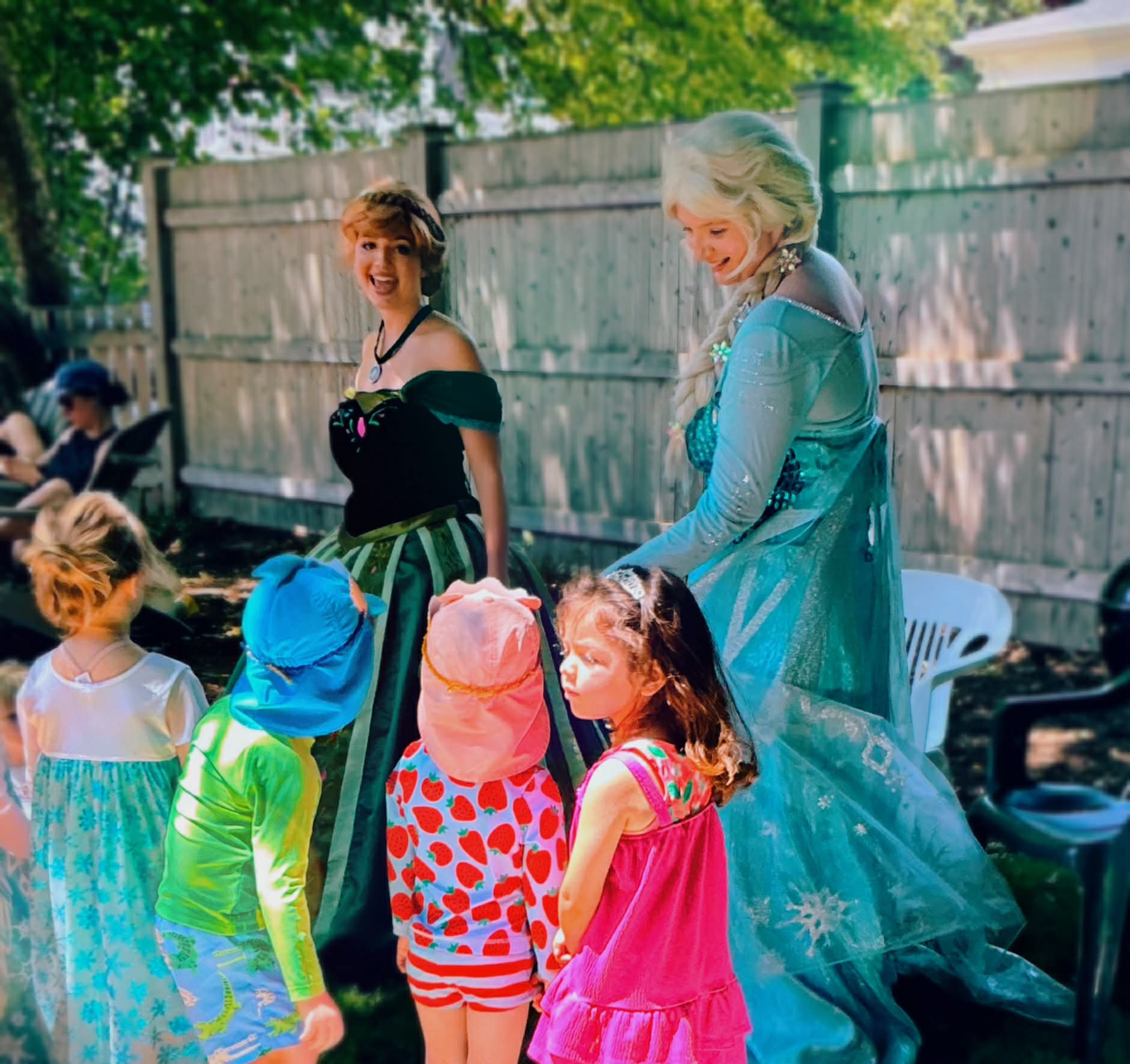 A group of children and the Snow Sisters party characters at a birthday party outdoors with a wooden fence and trees in the background.