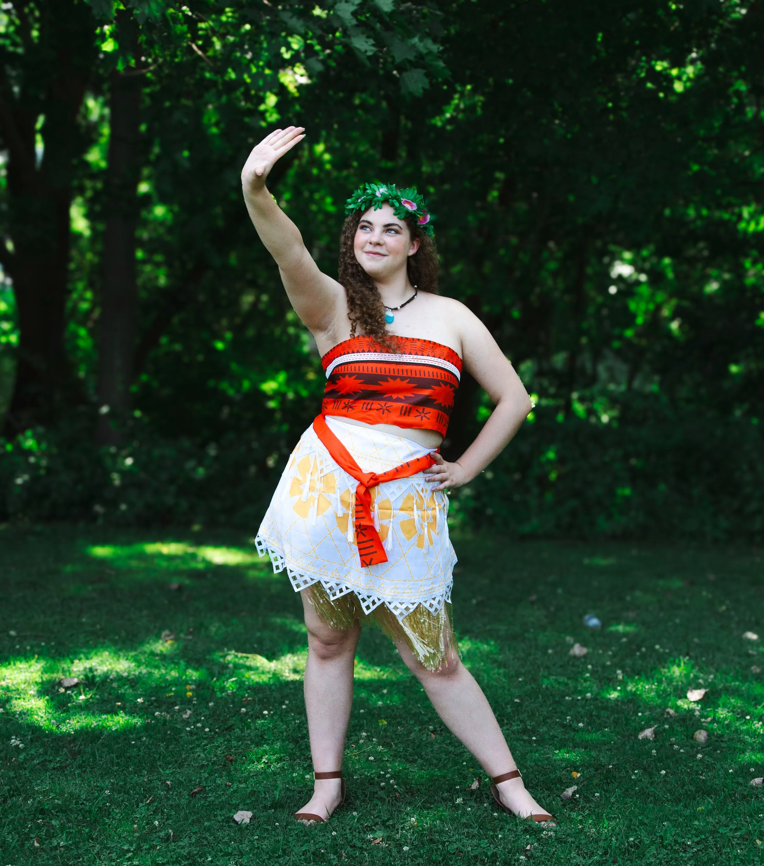 Island Voyager party princess dressed in a red, black, and white traditional Polynesian costume, wearing a leaf crown, standing in a green, wooded outdoor area, with one arm raised and a smile.