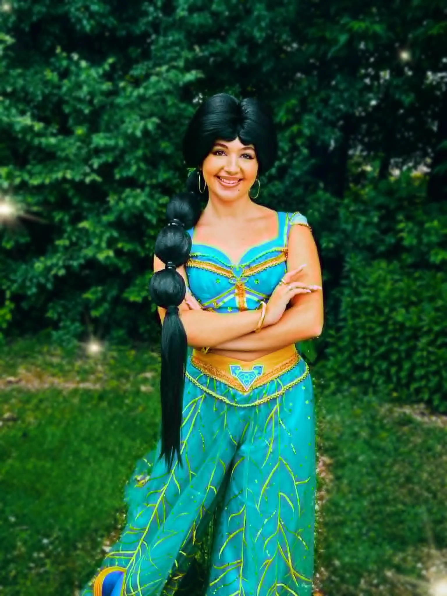 Arabian Princess Party Princess wearing a colorful dress with yellow and green details, stands outdoors in front of green foliage, smiling and crossing her arms.