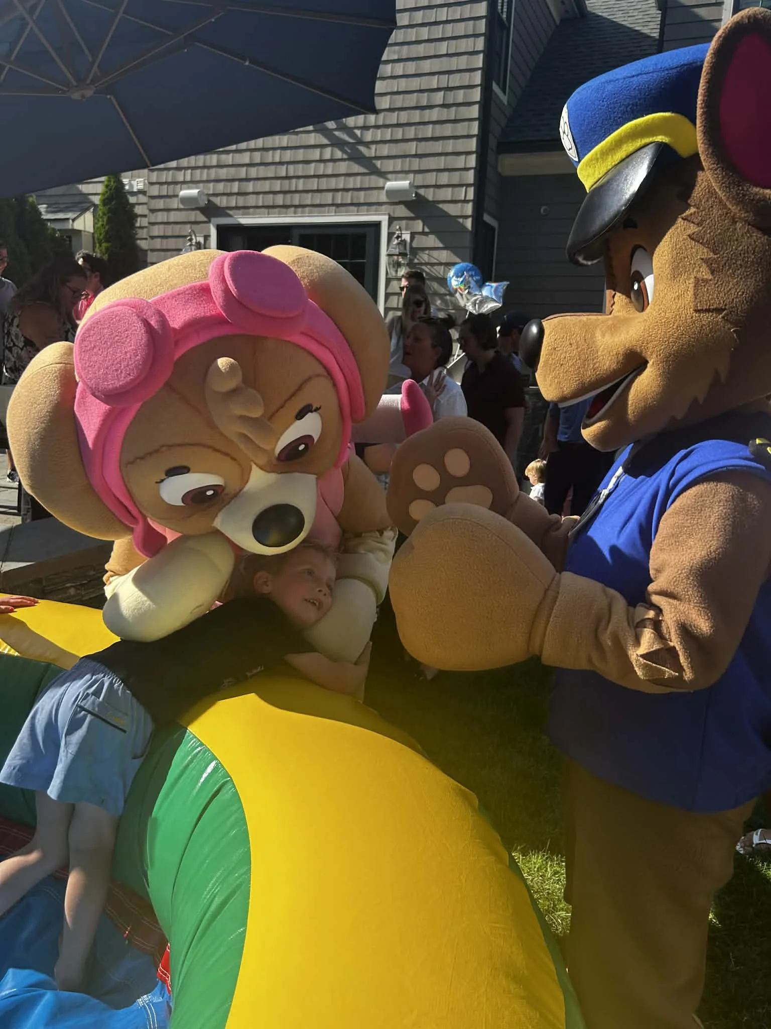 Children and adults playing with Patrol Pup party characters by colorful inflatable amusement park toy outdoors on a sunny day, with people and houses in the background.