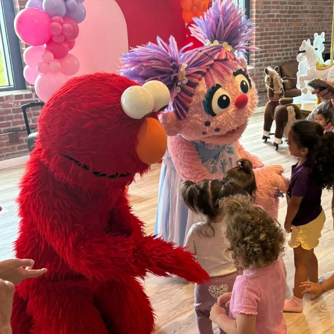 Children meeting Furry Red Monster and Fairy in Training party characters at a birthday party with pink and purple balloon decorations.