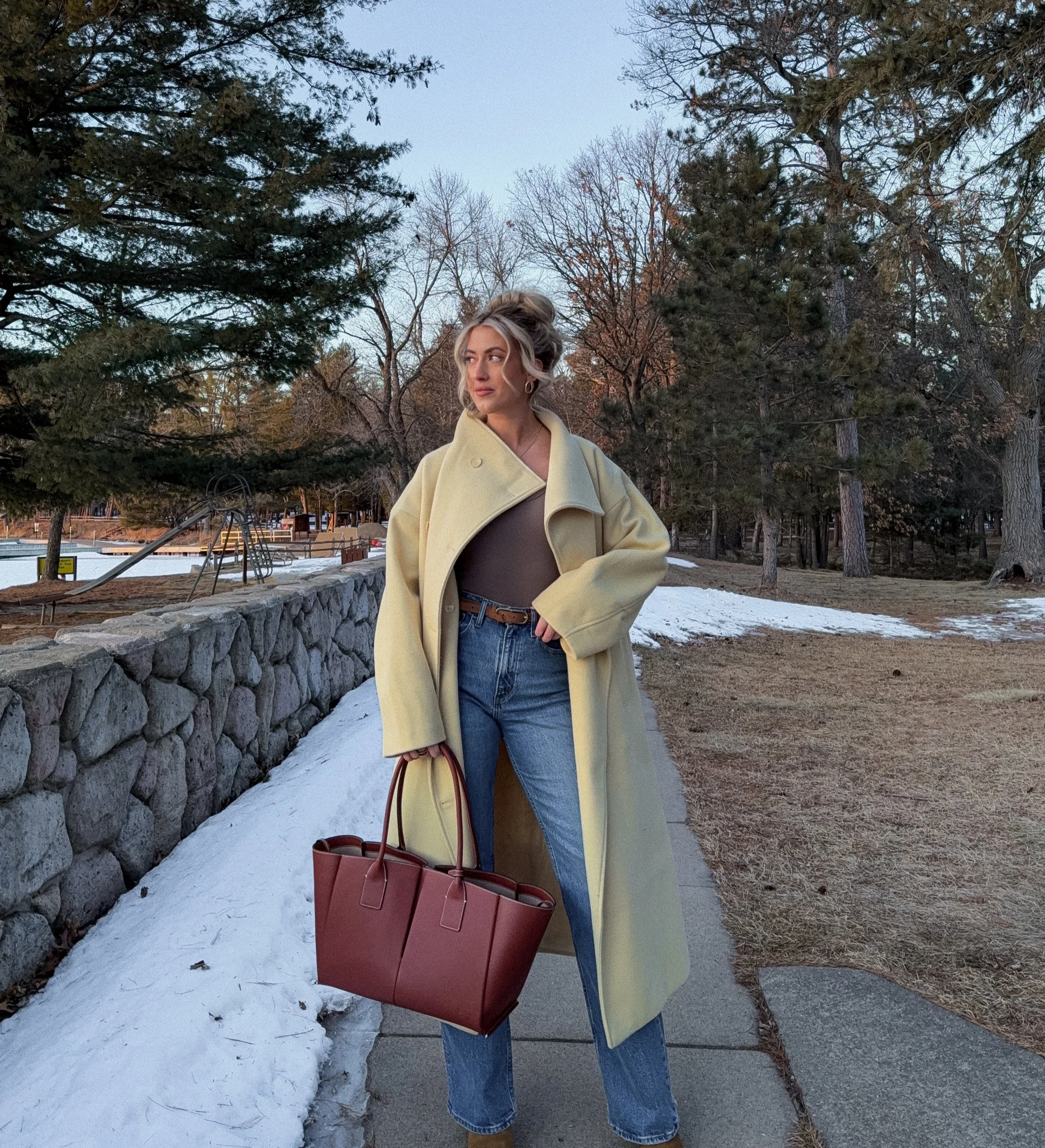 Woman standing on sidewalk in a park during late winter, wearing a yellow coat, brown top, jeans, and holding a large brown handbag, with trees and patches of snow in the background.