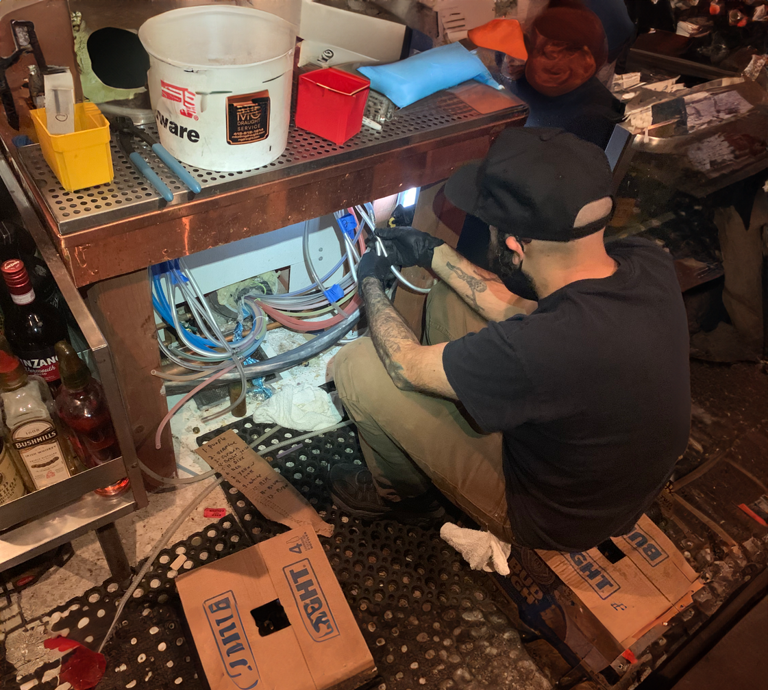 A person with tattoos, wearing a black t-shirt, khaki pants, a black cap, and gloves, sitting on the floor working on electrical or plumbing wires underneath a metal workbench in a bar or workshop. The area has various tools, cables, and boxes, with bottles of alcohol on a shelf nearby.