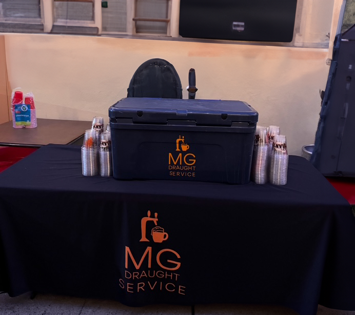 A black cooler on a table covered with a black tablecloth emblazoned with orange text and logo for MG Draught Service, accompanied by several stacks of clear plastic cups. In the background, there is a window, a chair, and cleaning supplies on a side table.