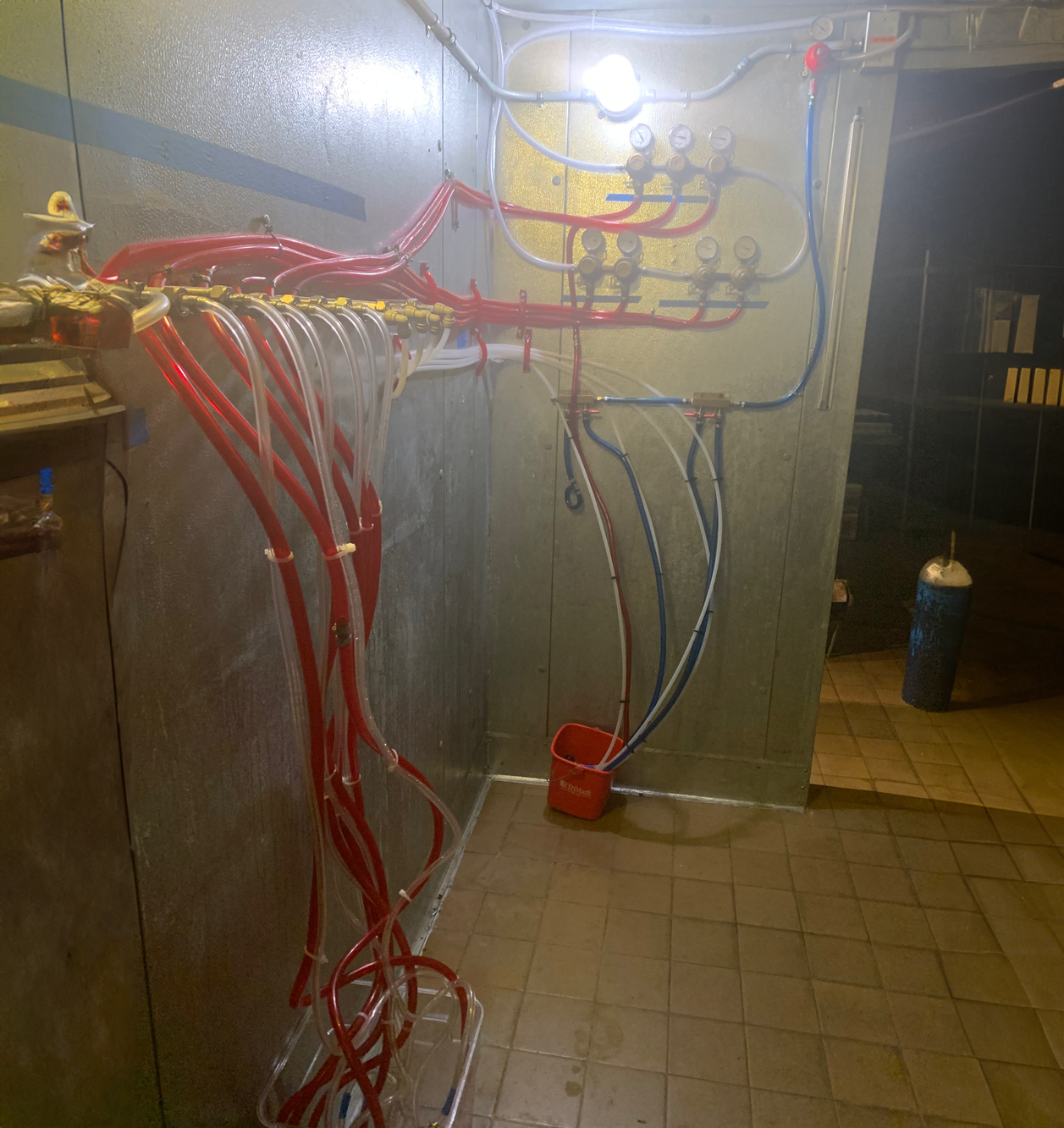 A mechanical or plumbing setup with numerous red and blue hoses connected to gauges and valves, mounted on a metal wall and floor. There is a red bucket with hoses on the floor and a large gas cylinder in the background.
