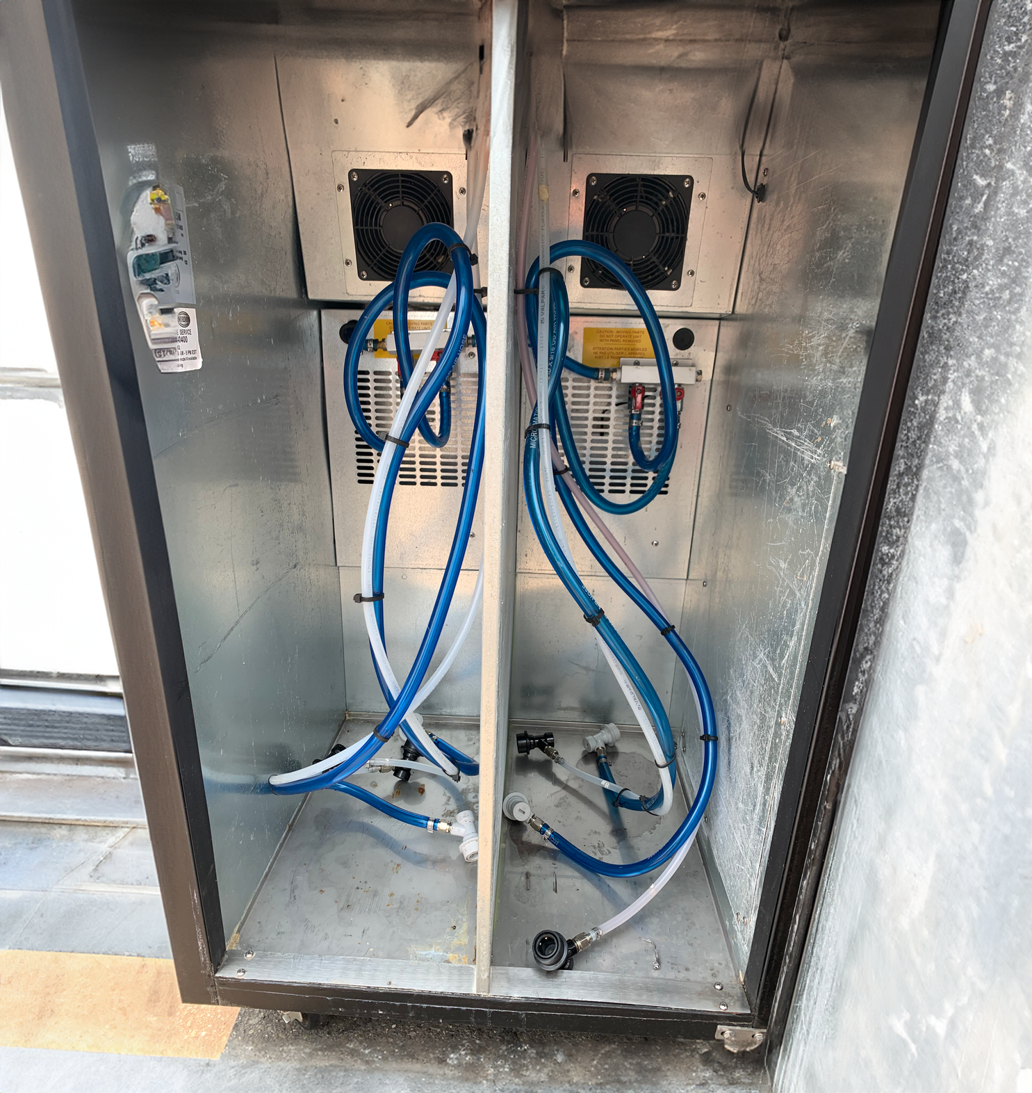 Inside a metal electrical or cooling box with blue and white tubes connected to the interior components and dual black fans installed on the back wall.