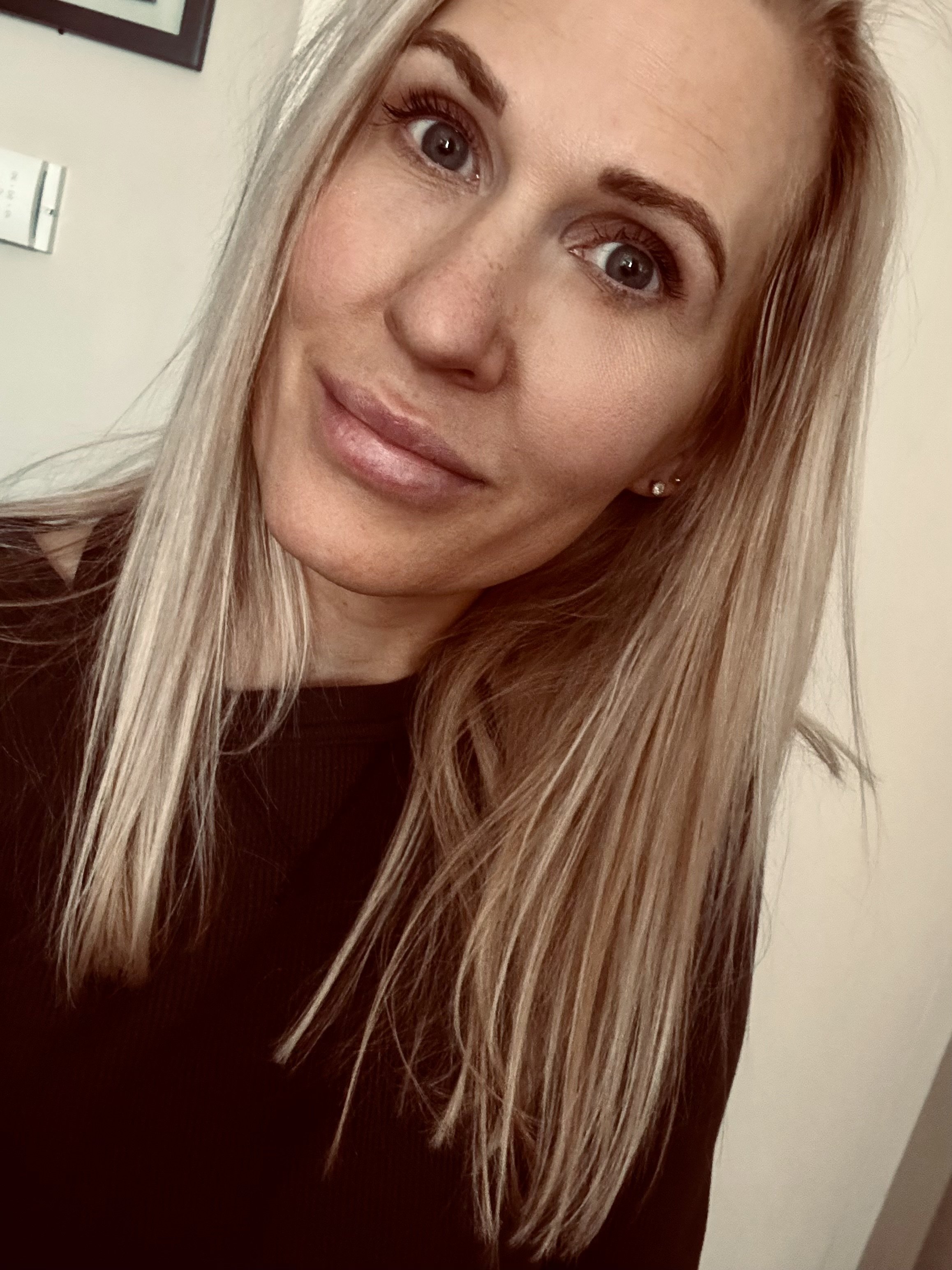 A close-up photo of a woman with long blonde hair, wearing makeup, earrings, and a dark top, smiling slightly and looking at the camera.