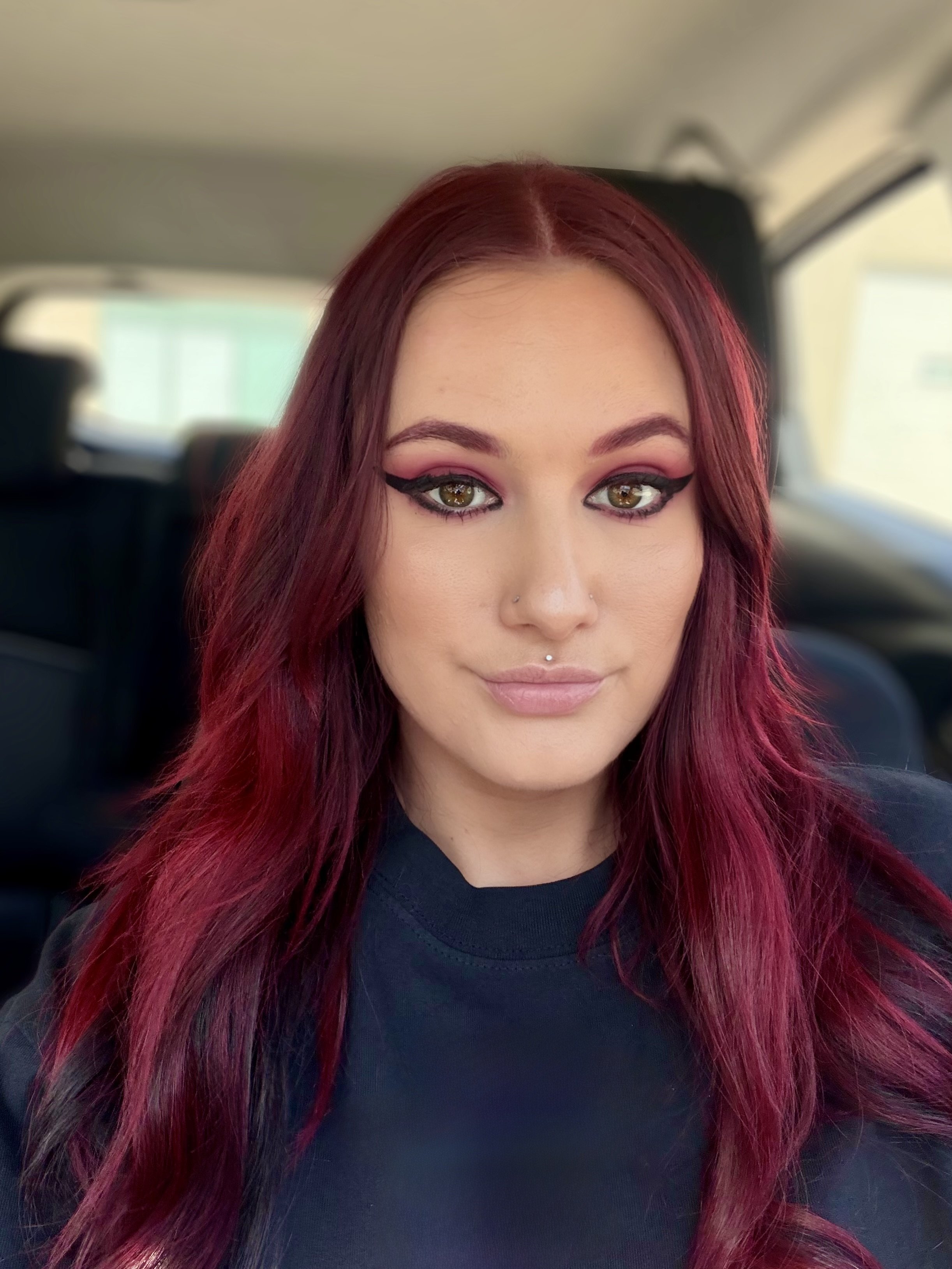 A close-up photo of a woman with long, burgundy hair and makeup, inside a vehicle.