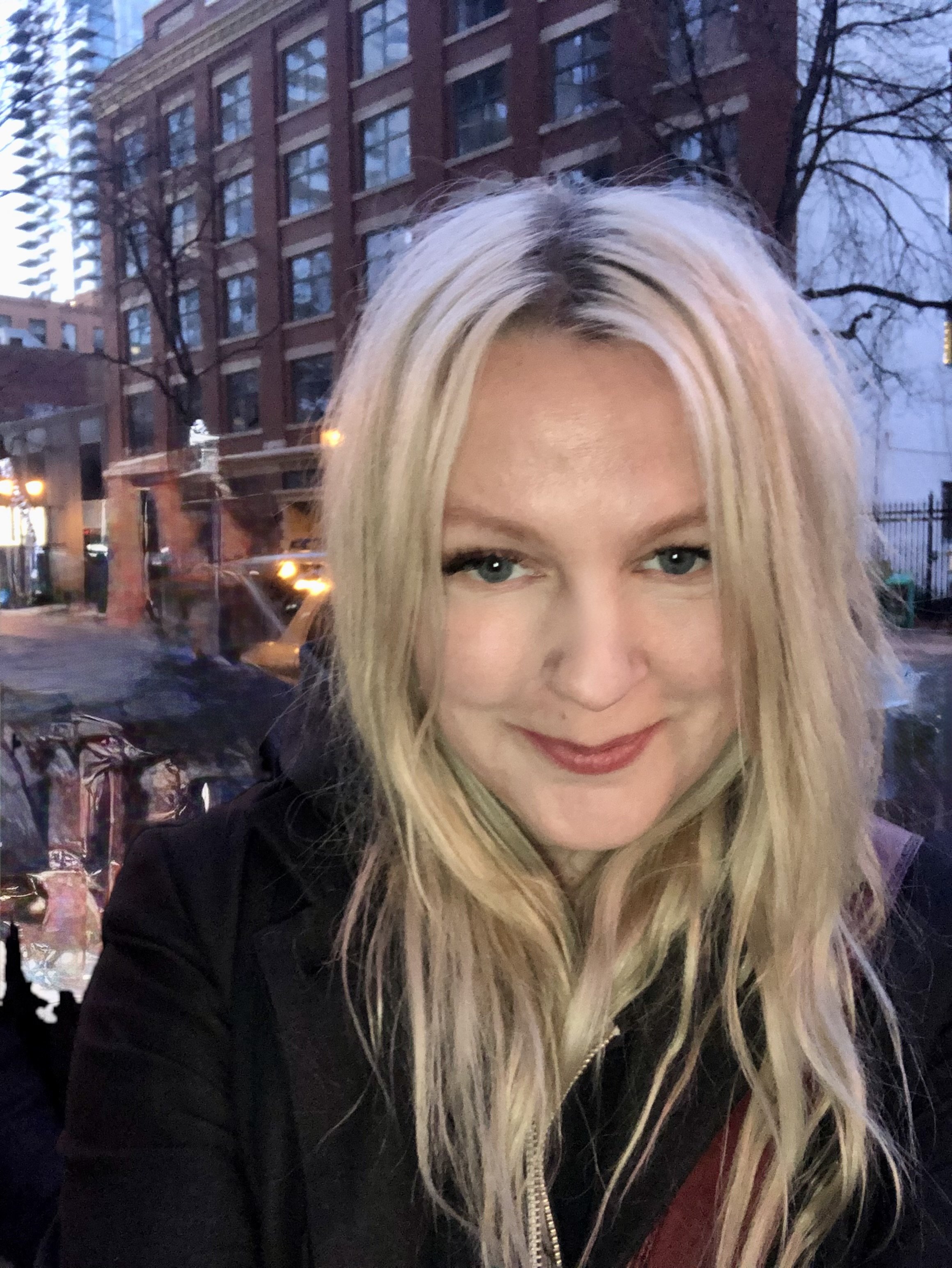 Close-up of a woman with long blonde hair and blue eyes, smiling, with city buildings and trees in the background during dusk or evening.