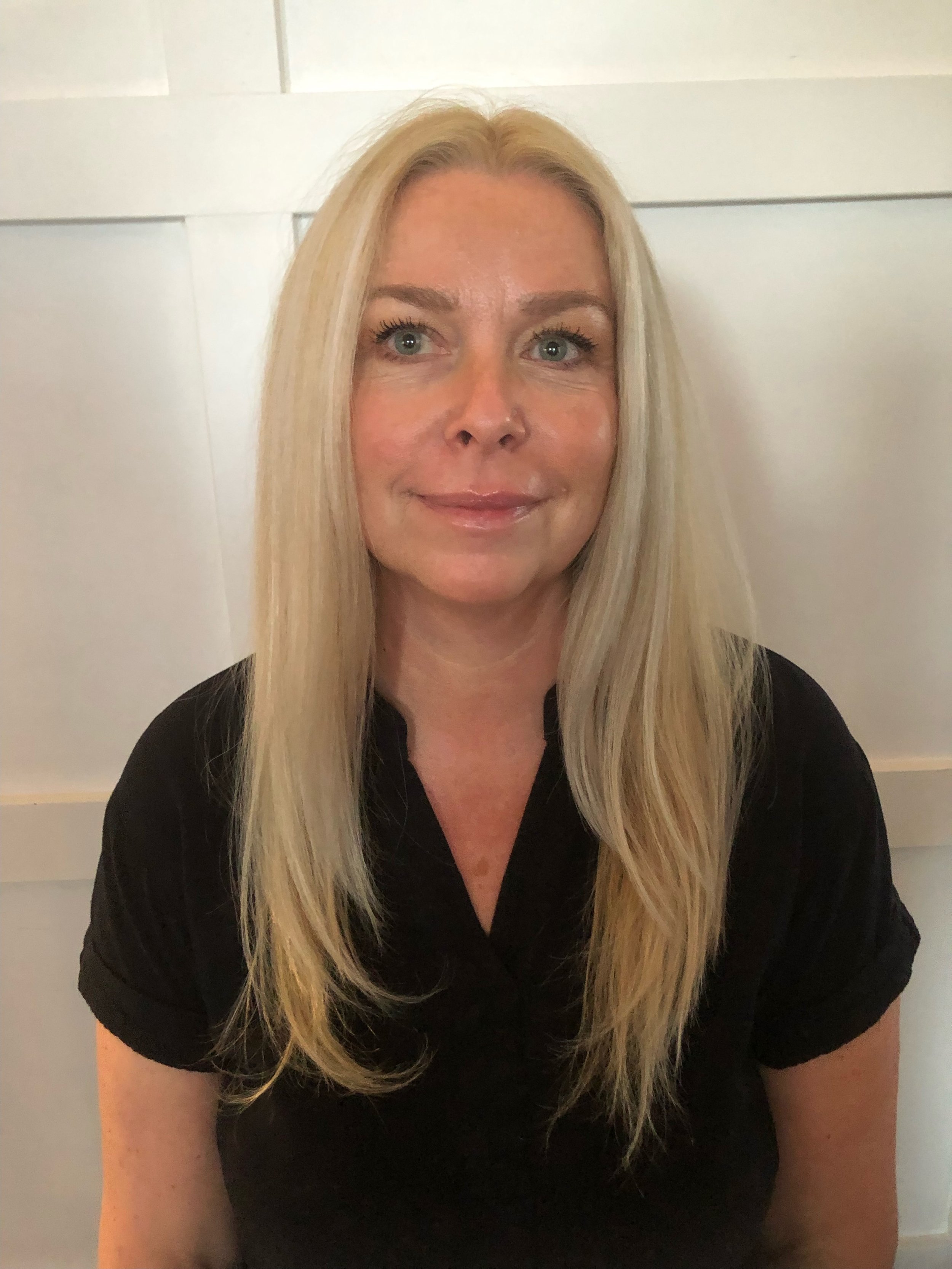 A woman with long blonde hair and blue eyes, smiling slightly, wearing a black top, sitting in front of a light-colored wall with horizontal panels.