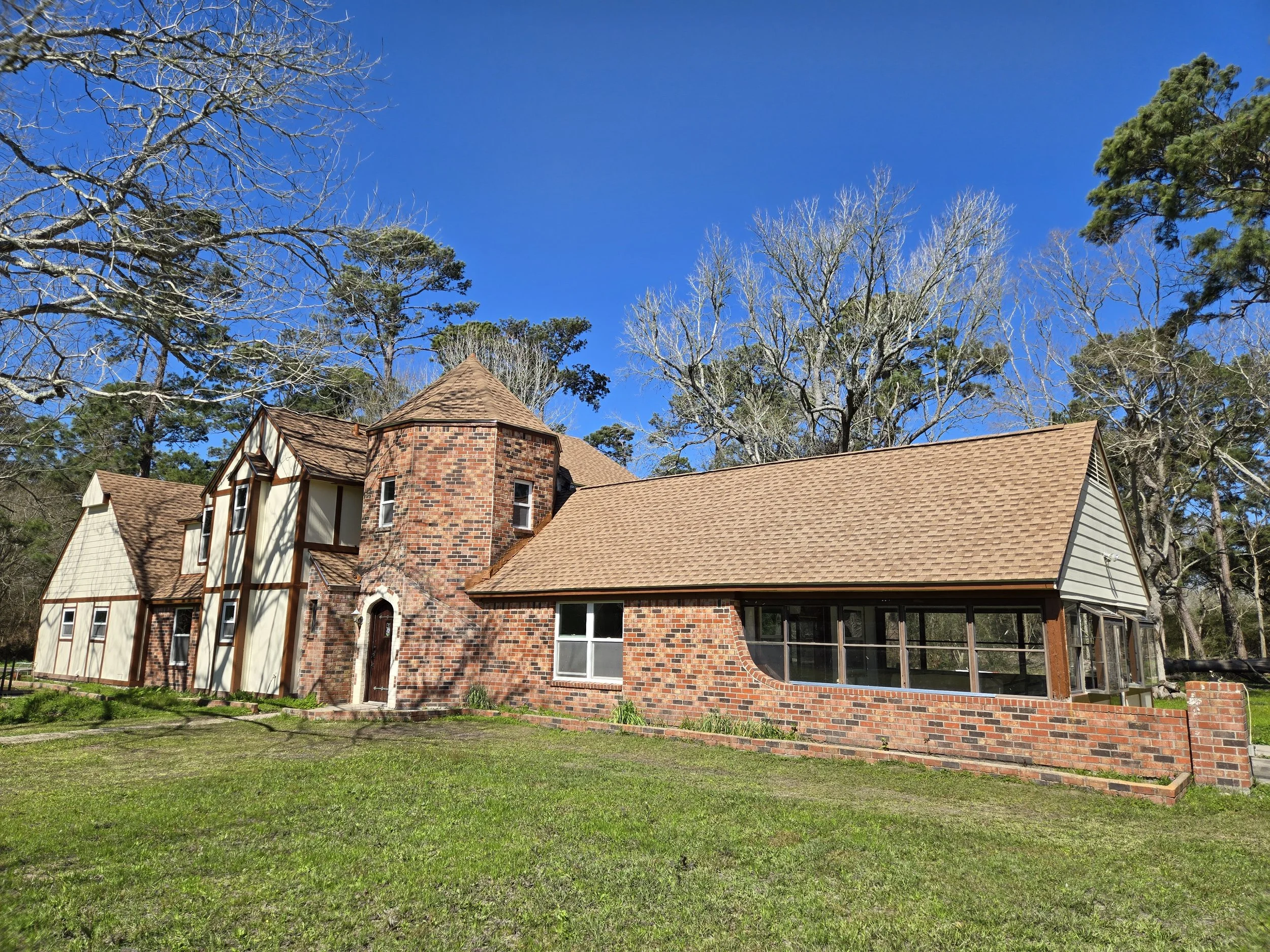A large house with a brick and wood exterior, multiple gabled roofs, and a tall brick tower. The house is surrounded by trees and a grassy lawn, with a clear blue sky overhead.
