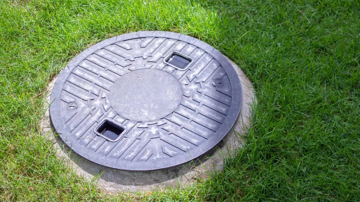 A round black manhole cover with two square handles, set in a concrete surround, surrounded by green grass.