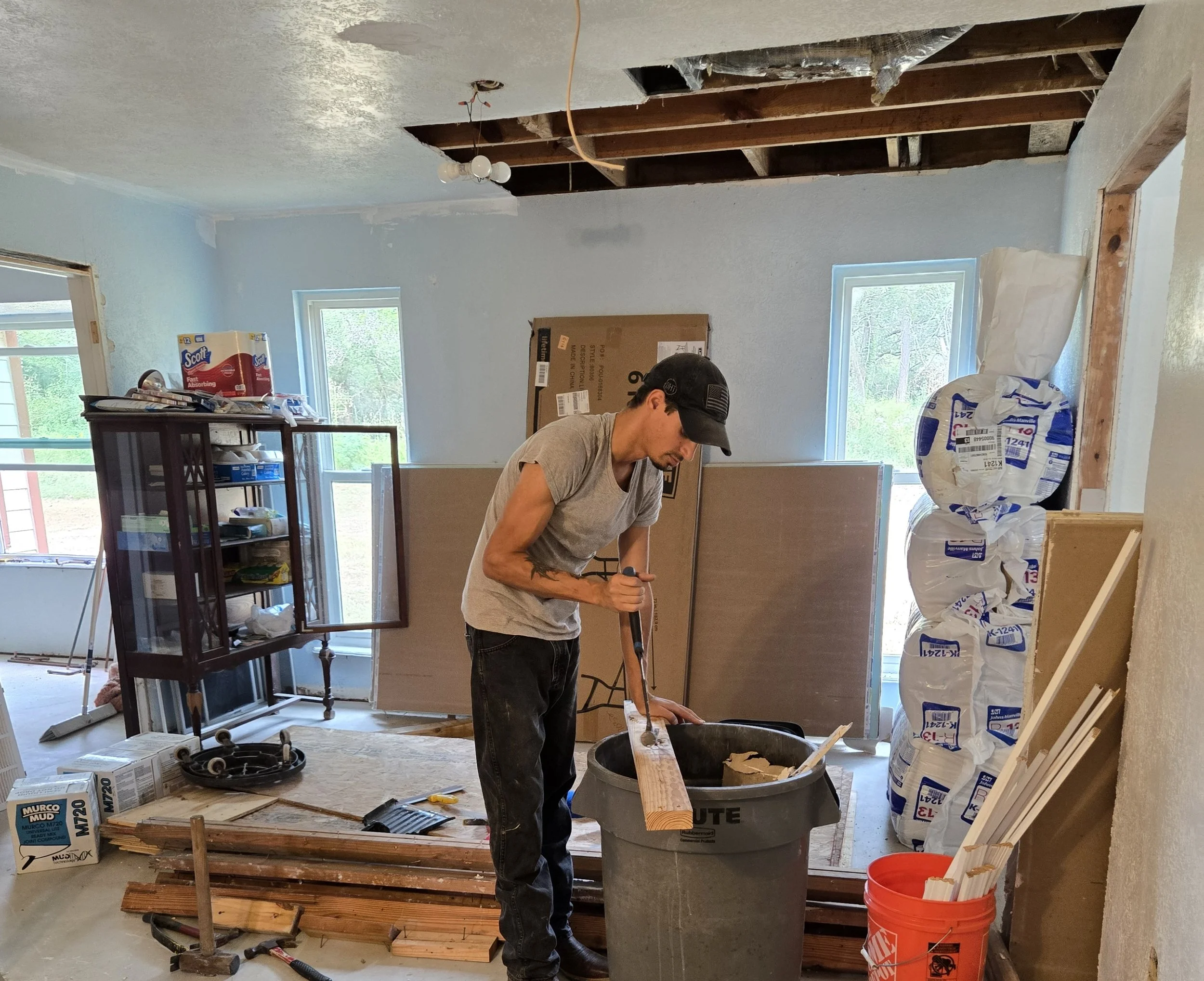 A man in a gray shirt and black cap is working inside a room under construction. He is using a tool with a long handle, possibly a crowbar, to work on a piece of wood over a gray trash bin. The room has insulation bags stacked on the right, a shelving unit on the left, and partially finished walls with some drywall and exposed ceiling beams.