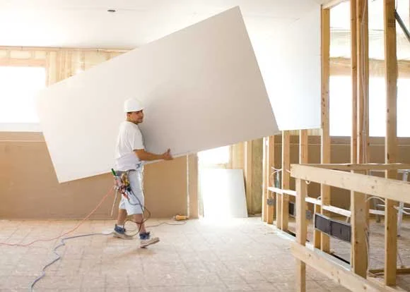 Construction worker carrying a large white drywall panel inside a building under construction.
