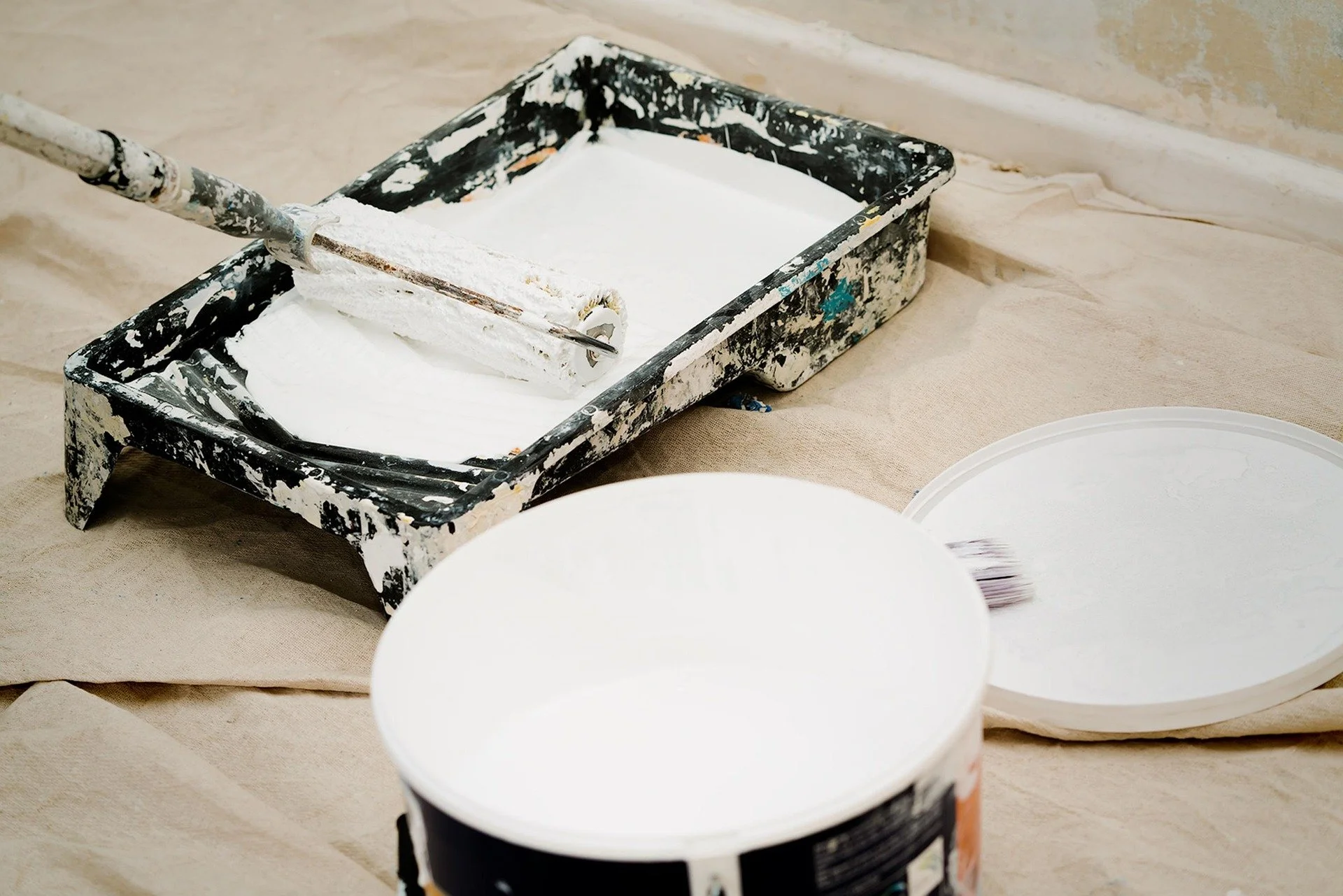 Paint roller dipped in white wall paint resting on a paint tray filled with white paint, with empty paint cans and lids nearby on a tan-colored protective covering.