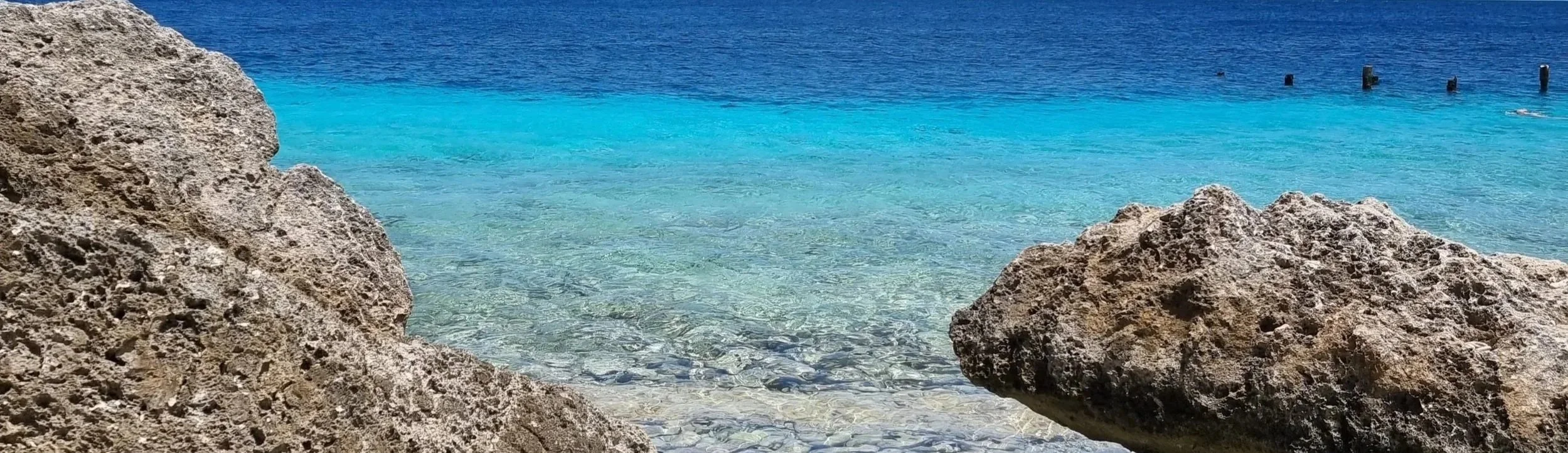Clear blue ocean water with rocks in the foreground and weathered wooden posts in the distance.