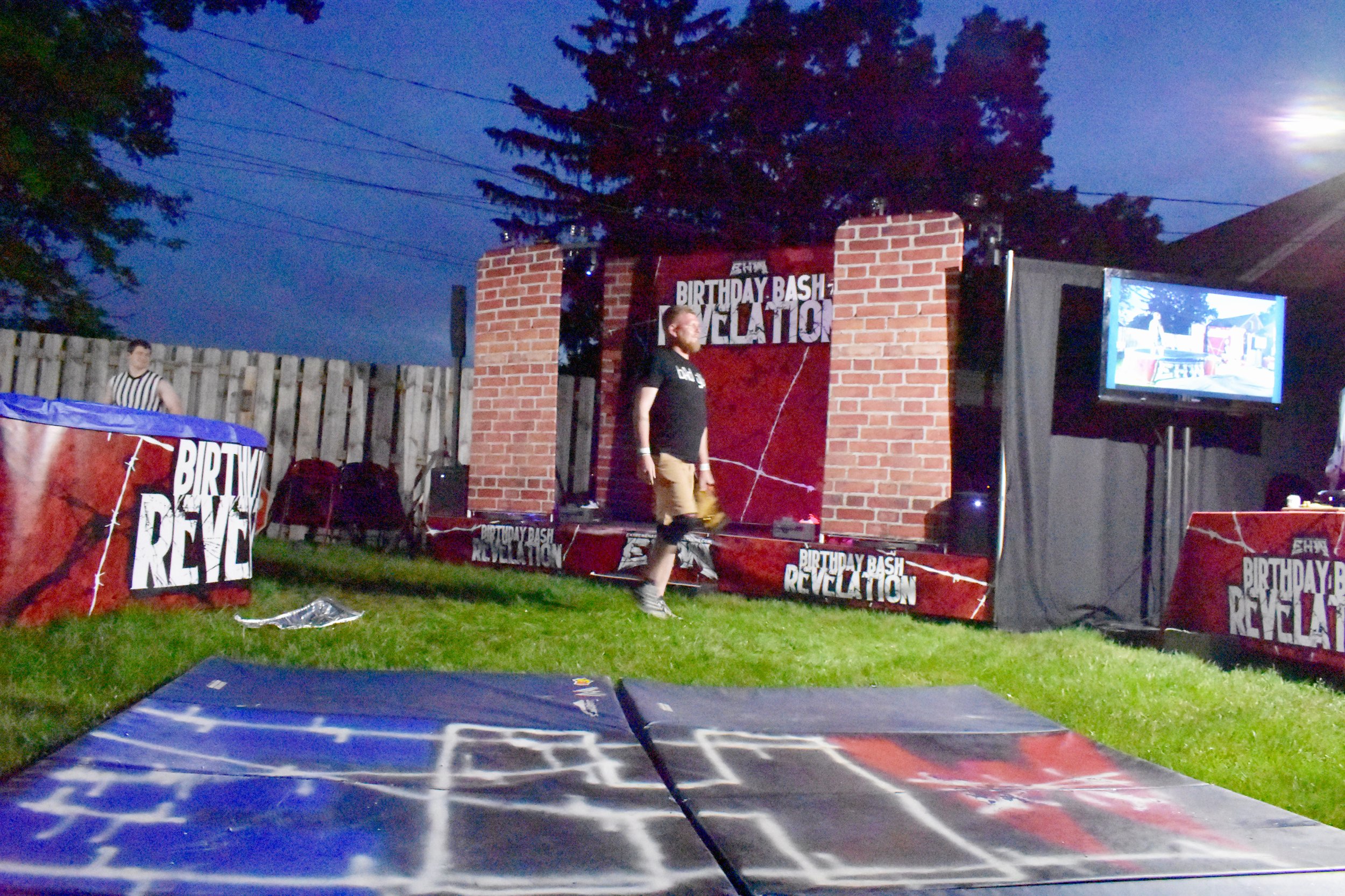 Outdoor wrestling event setup with a stage labeled "Birthday Bash Revelation," featuring a person walking, a referee, decorated panels, and a screen at dusk.