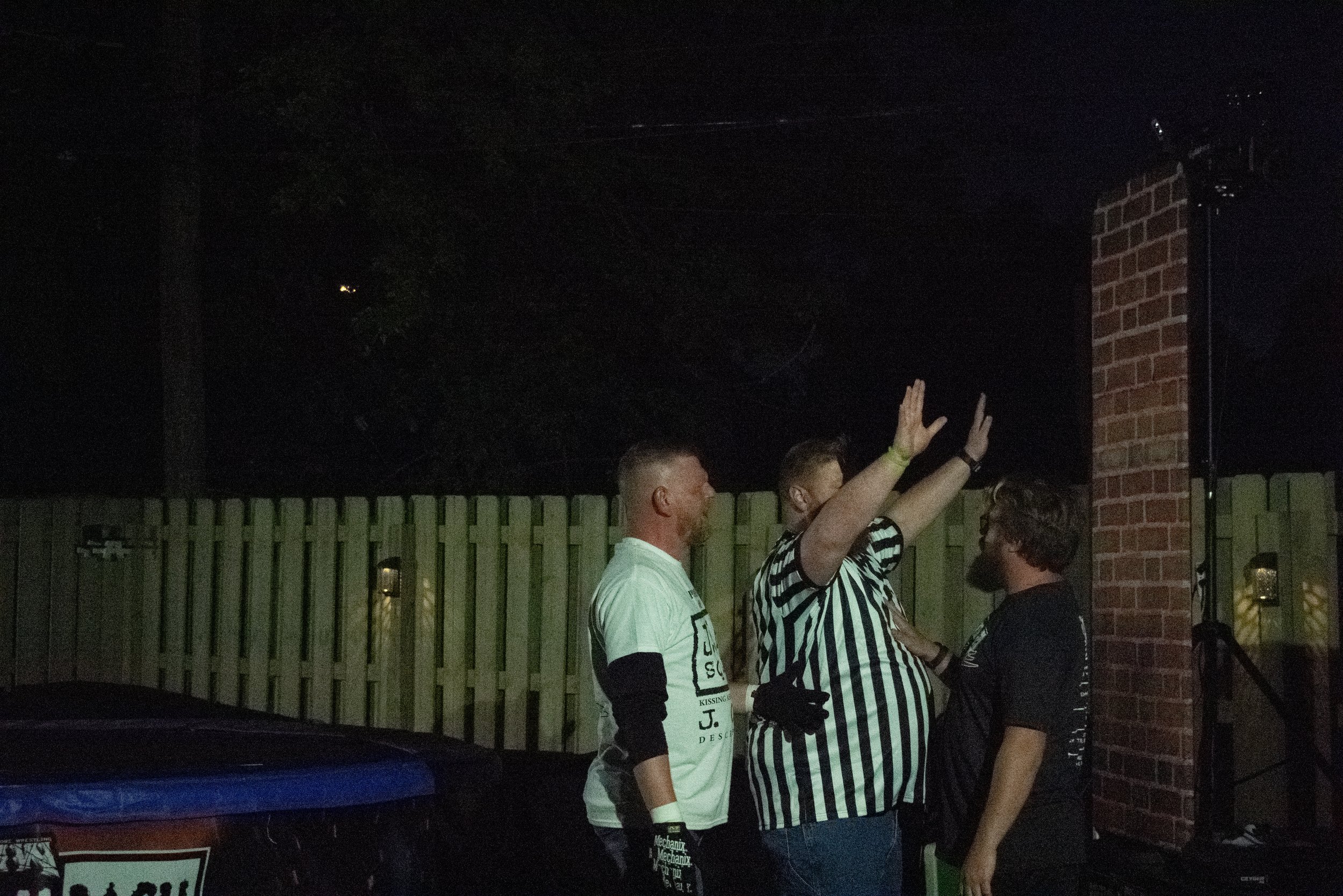 Three men outside at night, one wearing a referee shirt, appear to be engaged in a conversation or argument, near a wooden fence.