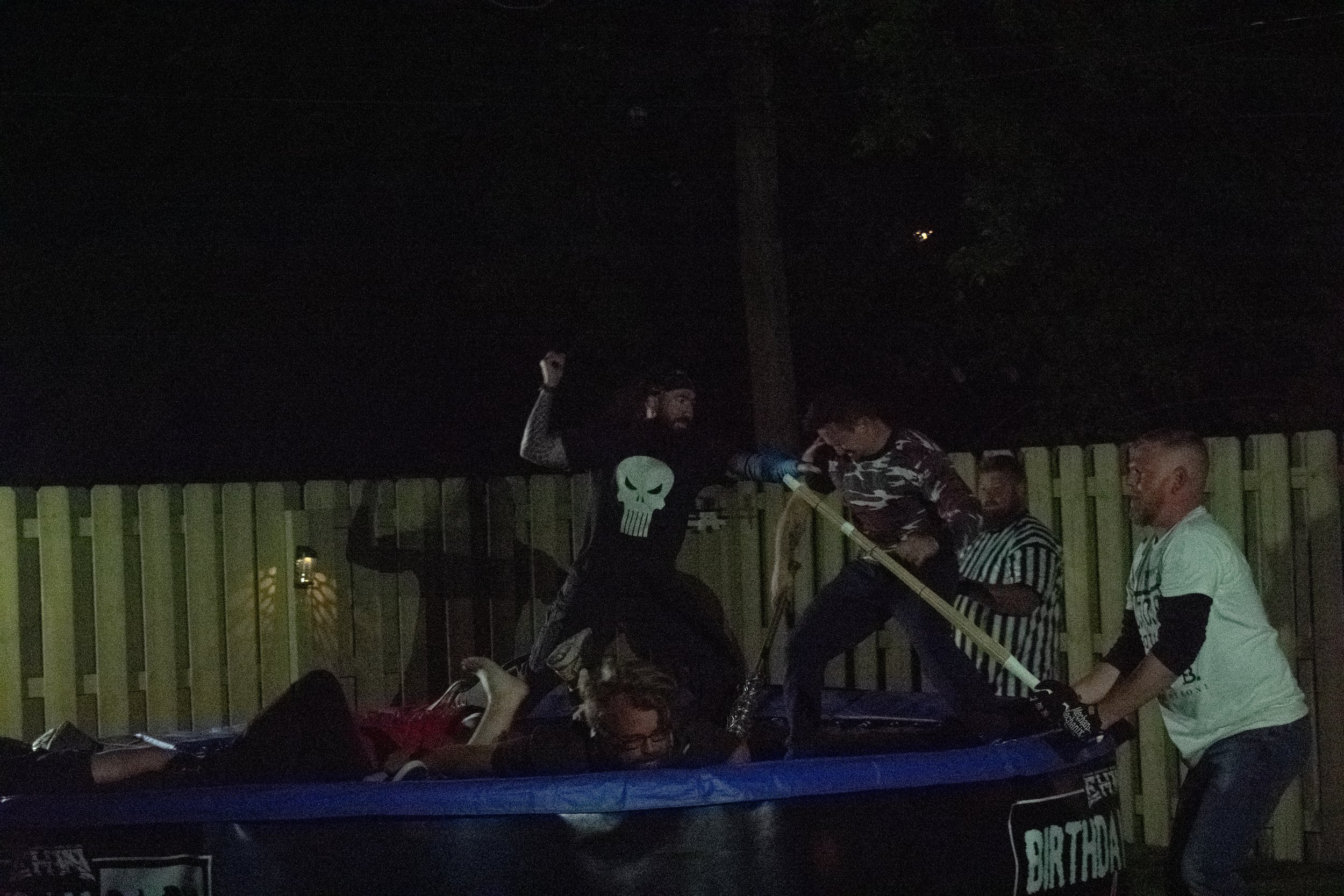 Group of people in a wrestling match on a trampoline at night, surrounded by a wooden fence. One participant is wearing a skull emblem shirt.