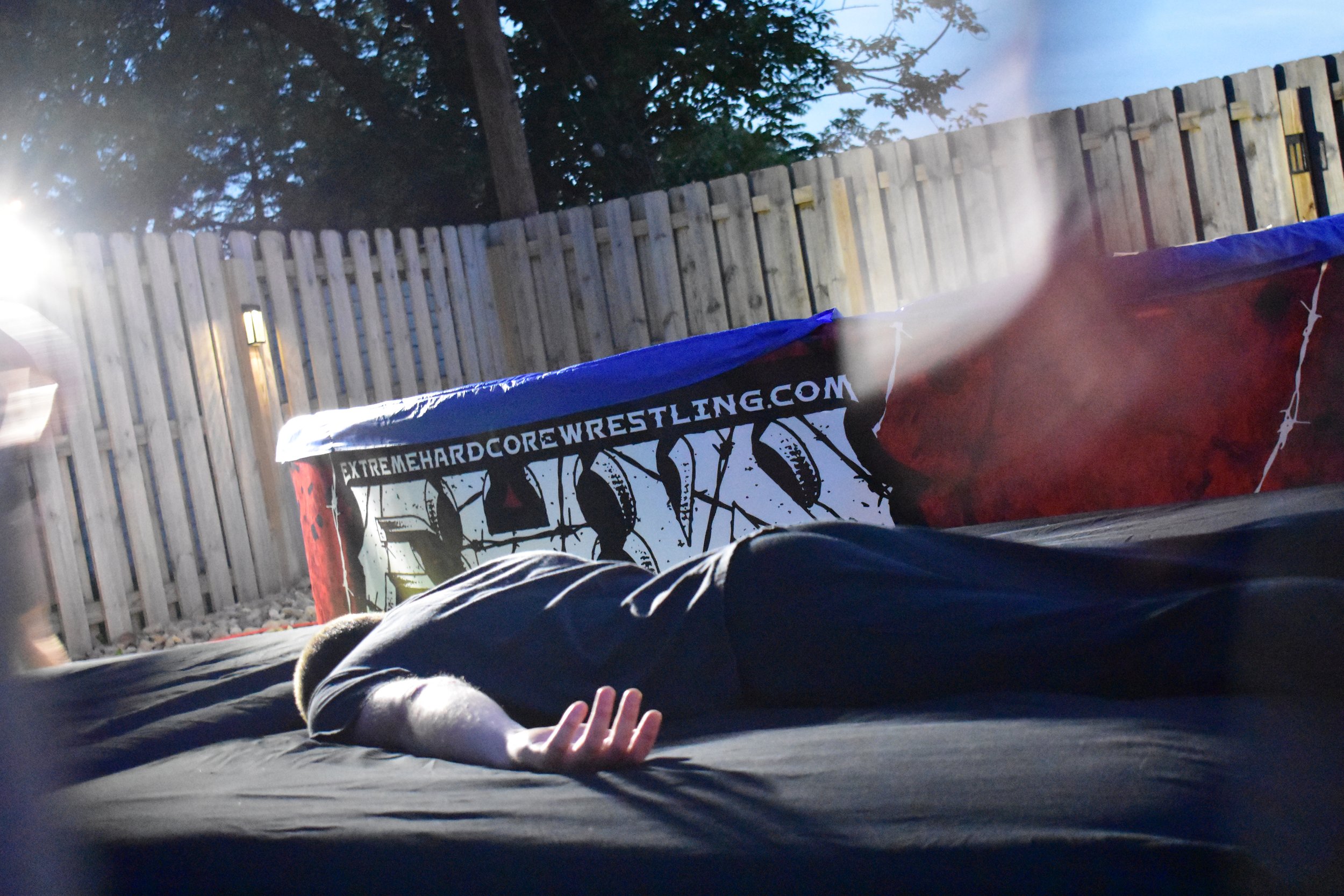 Outdoor wrestling ring setup with person lying on mat, surrounded by wooden fence, banner displays "extremehardcorewrestling.com."