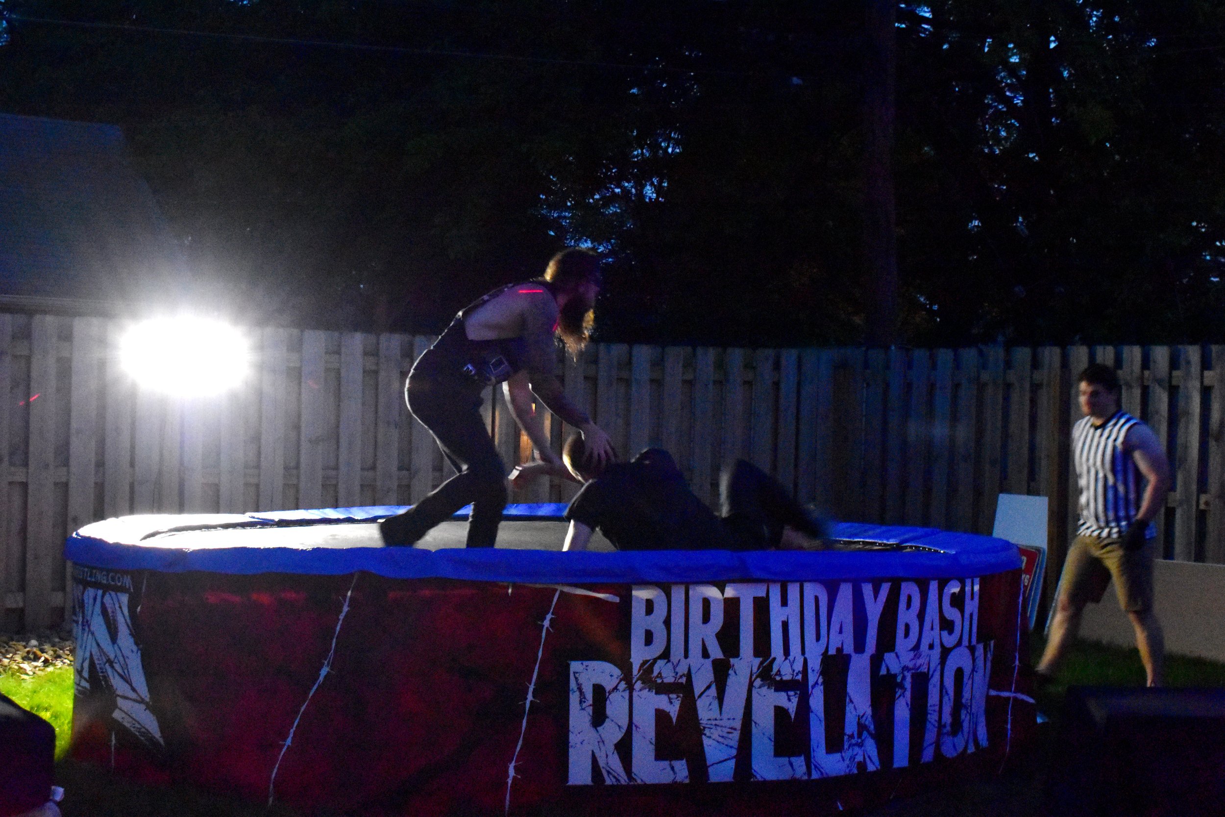 Outdoor wrestling match in progress with two participants on a trampoline labeled 'Birthday Bash Revelation.' A referee is seen on the right.