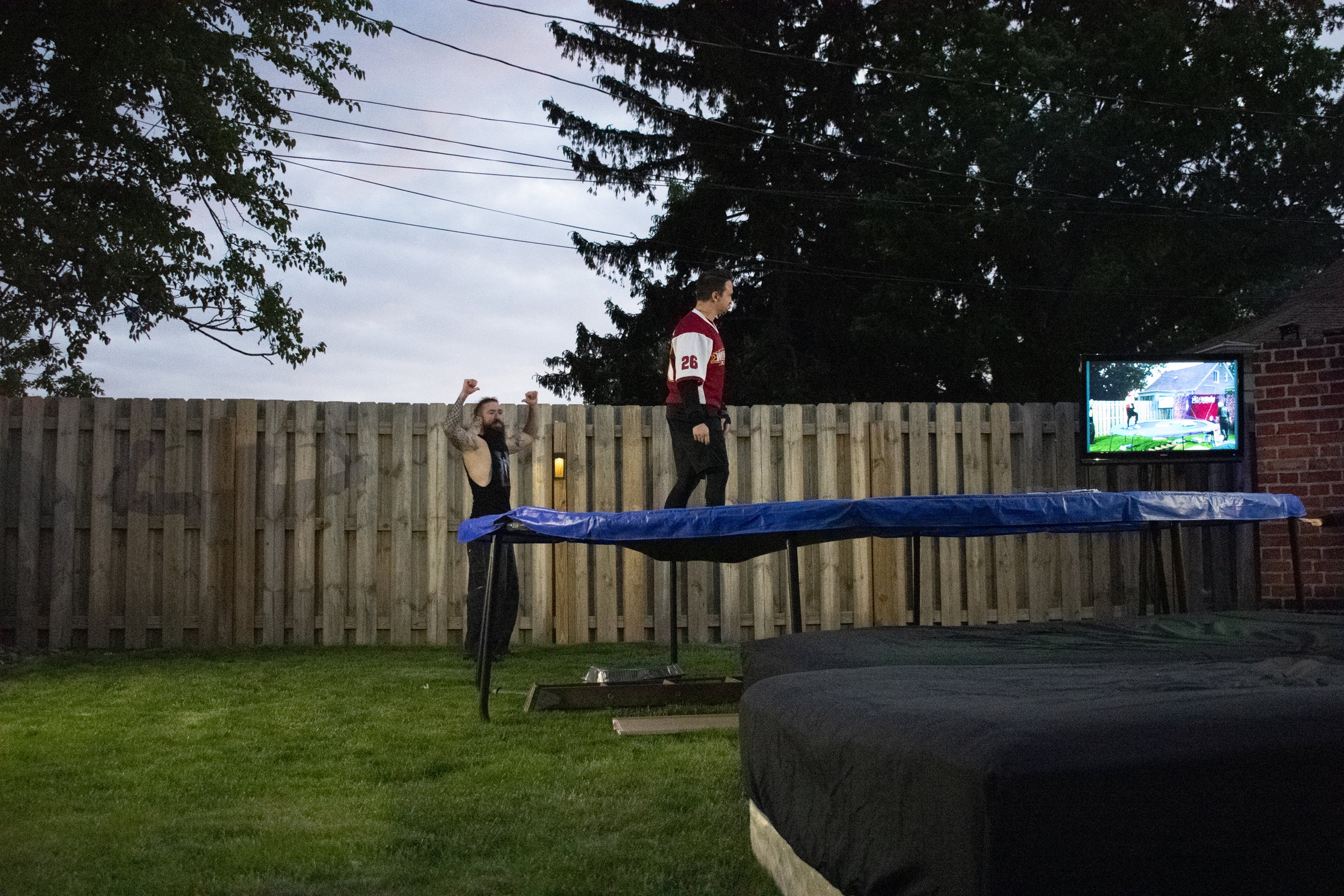 Two people in a backyard wrestling setup, one standing on a trampoline, the other raising arms in celebration, with a wooden fence and trees in the background.
