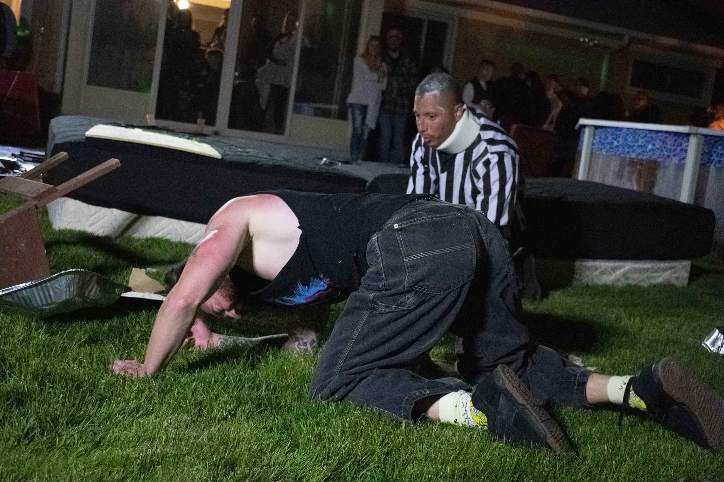 A person kneeling on grass, with a referee in a striped shirt and neck brace observing. In the background, there are people watching, and a broken chair is on the ground beside an overturned tray.
