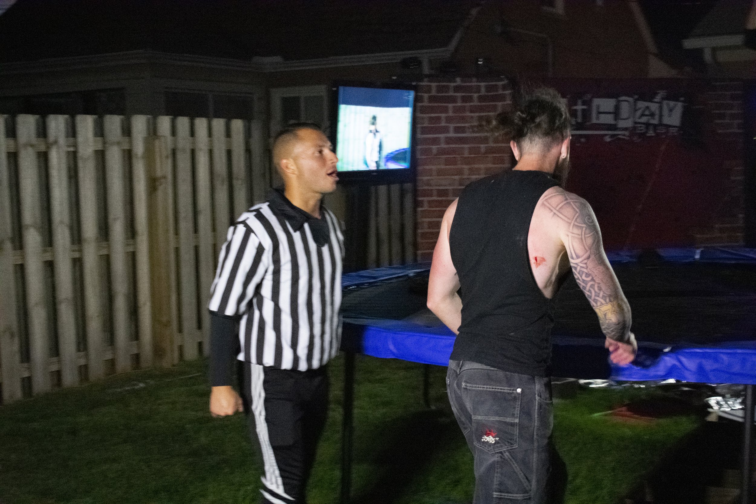 Referee and tattooed man near a backyard trampoline at night, with a screen and wooden fence in the background.