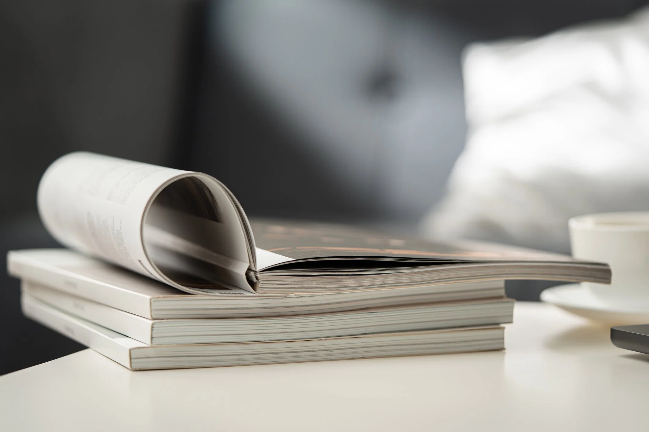 A stack of white books on a white table, with one open book placed on top of the stack, and a blurred background with a cup and other objects.