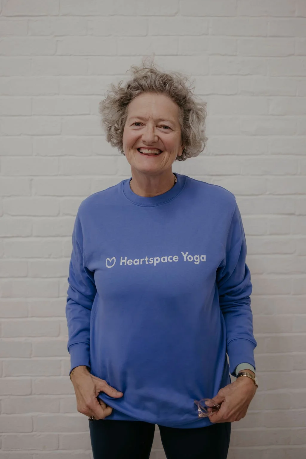 A woman with curly gray hair smiling, wearing a blue sweatshirt with 'Heartspace Yoga' written on it, standing in front of a white brick wall.