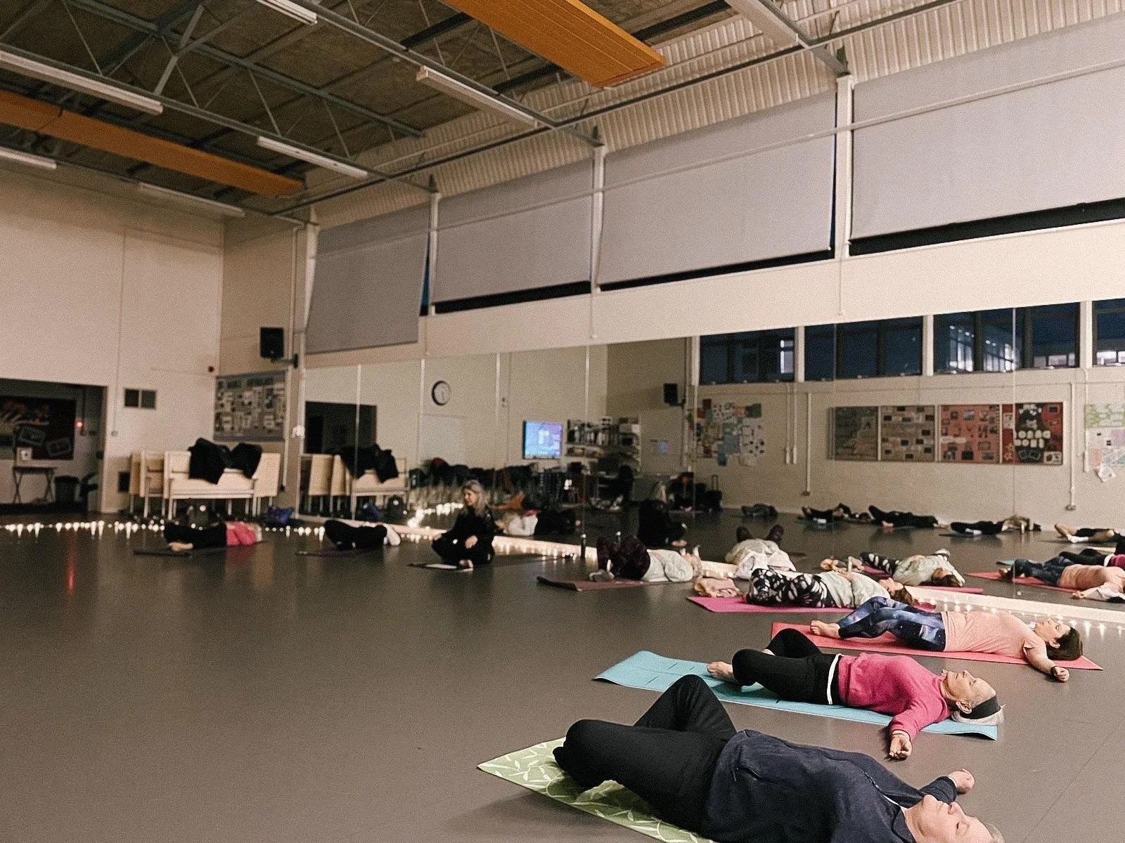 People participating in a yoga class or meditation session on mats in a spacious indoor studio with high ceilings, large windows, and wall-mounted artwork.