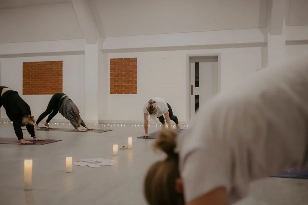 Group of people doing yoga in a dimly lit room with candles along the floor, brick accents on the wall, and a door in the background.