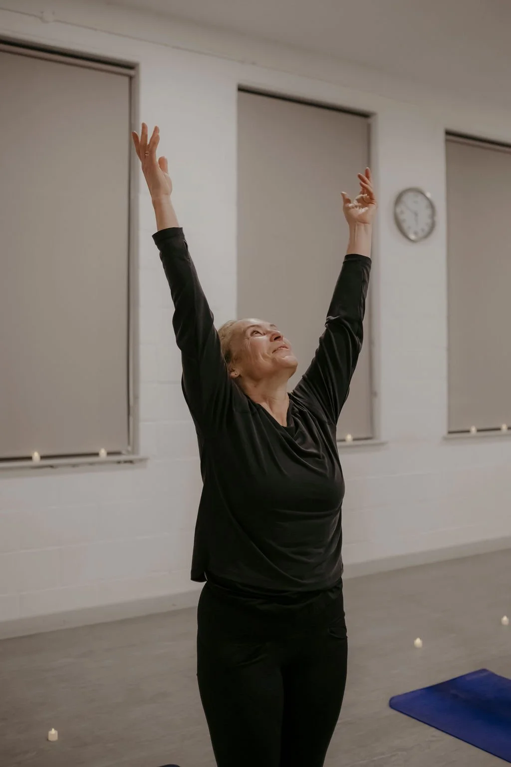 An elderly woman practicing yoga or stretching with arms raised in a spacious indoor room, with windows and a clock on the wall.
