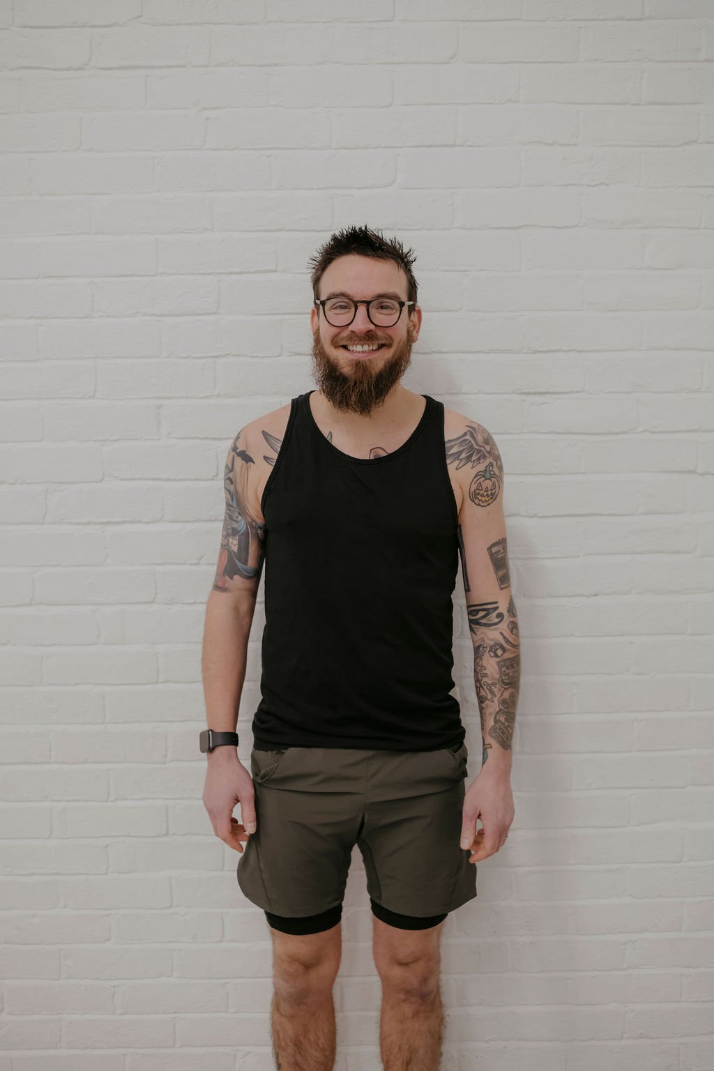 A smiling man with a beard and glasses standing against a white brick wall, wearing a black tank top and brown shorts.