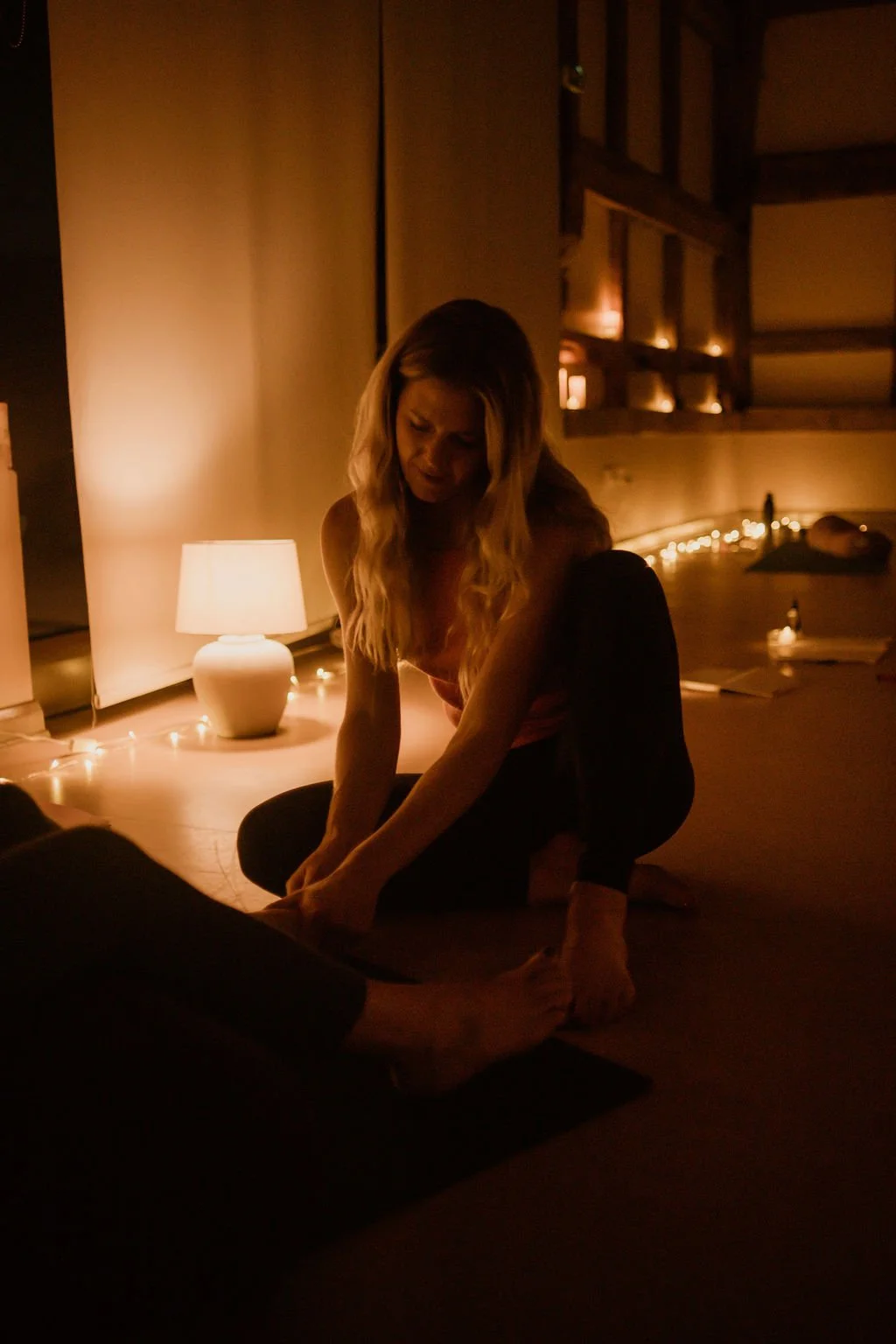 A woman practicing yoga or meditation on a mat in a softly lit room with warm lighting, a desk lamp, and string lights in the background.