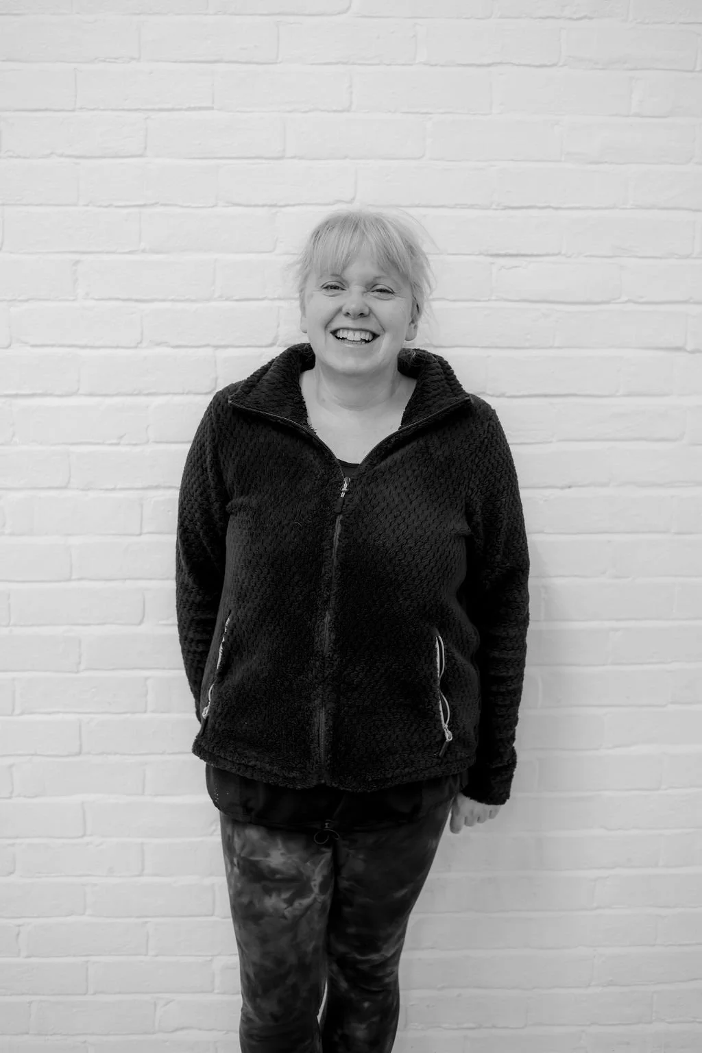 A smiling woman with short light-colored hair standing against a white brick wall, wearing a black zip-up fleece jacket and patterned pants.