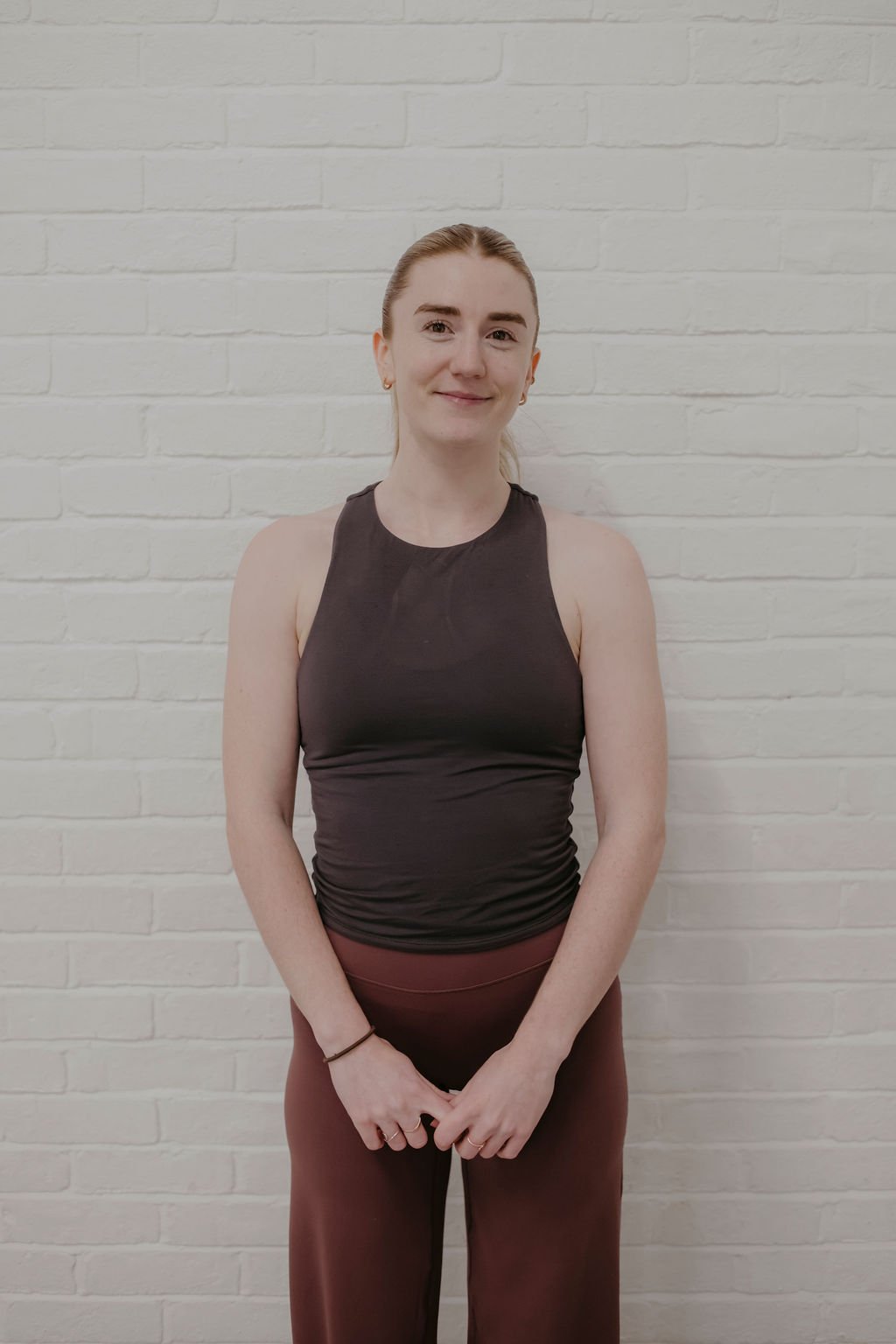 A young woman with light skin, reddish hair tied back, wearing a black sleeveless top and maroon pants, standing against a white brick wall, smiling gently.