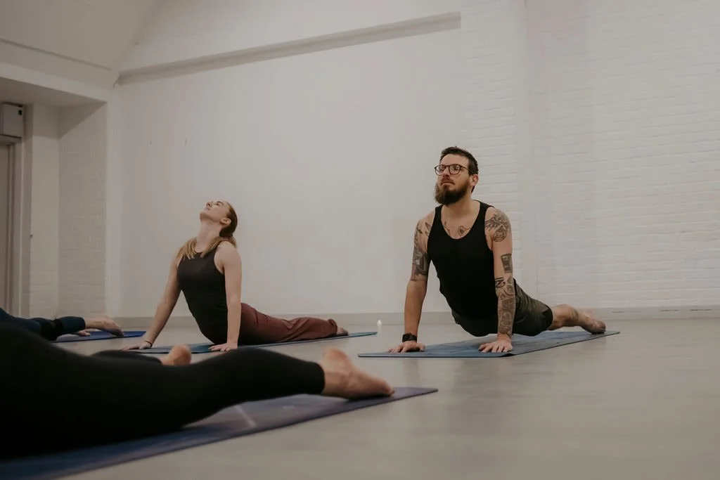 A man with tattoos and glasses in a black tank top is practicing yoga on a mat in a studio, with a woman in a black tank top and maroon pants doing a similar pose nearby.