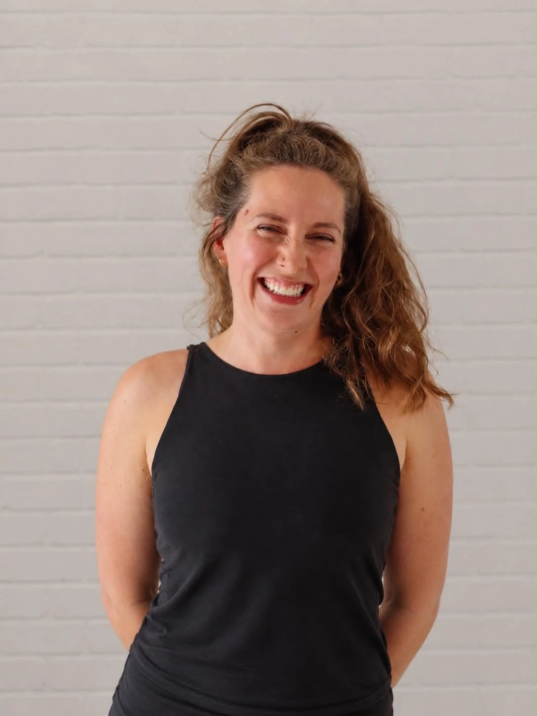 A woman with curly brown hair in a high ponytail, wearing a black sleeveless top, smiling and standing in front of a white brick wall.