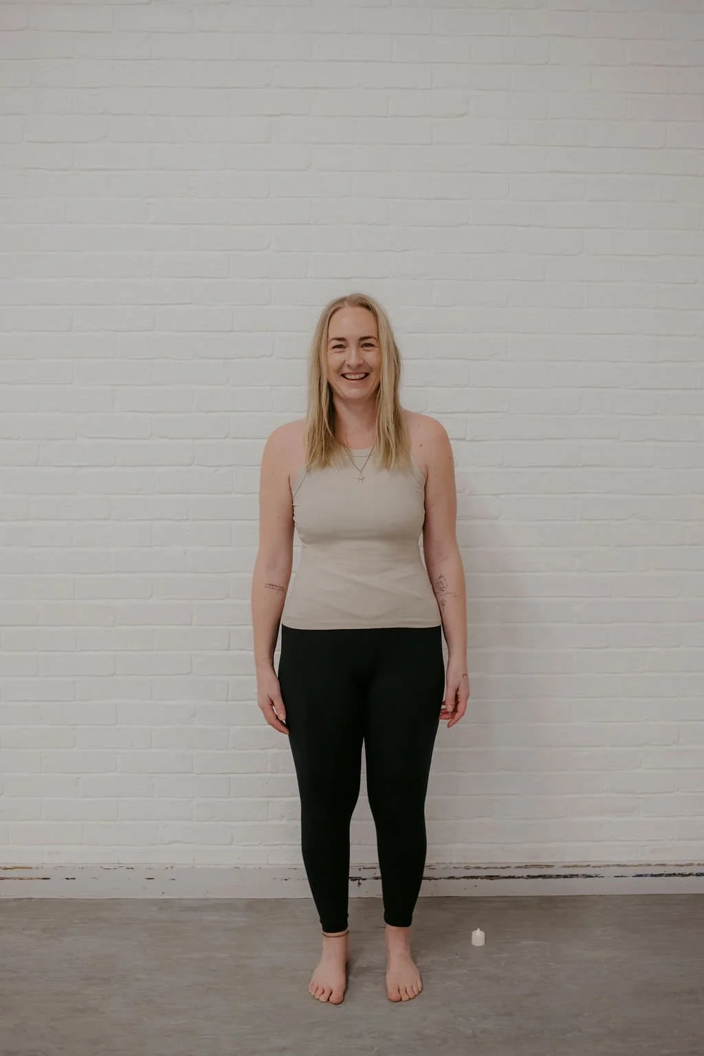 A woman with long blonde hair standing barefoot against a white brick wall, wearing a beige sleeveless top and black pants, smiling at the camera.