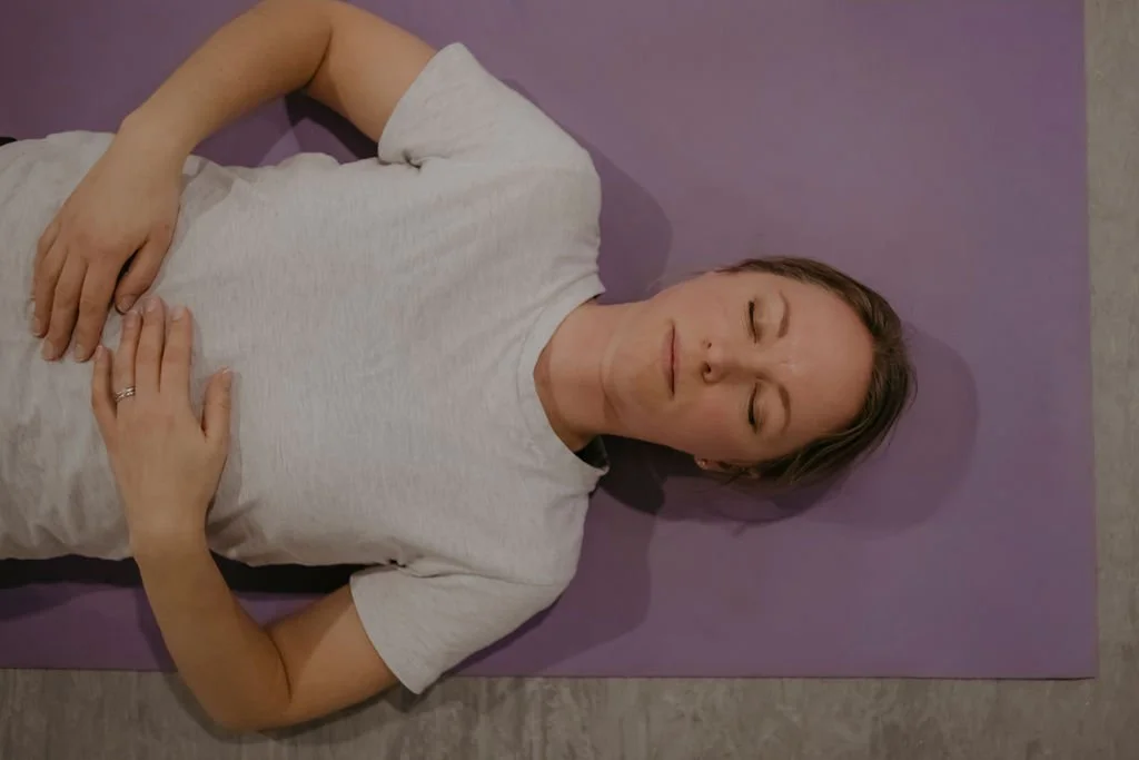 A woman lying on her back on a purple yoga mat with her eyes closed, resting with one hand on her chest and the other on her stomach.