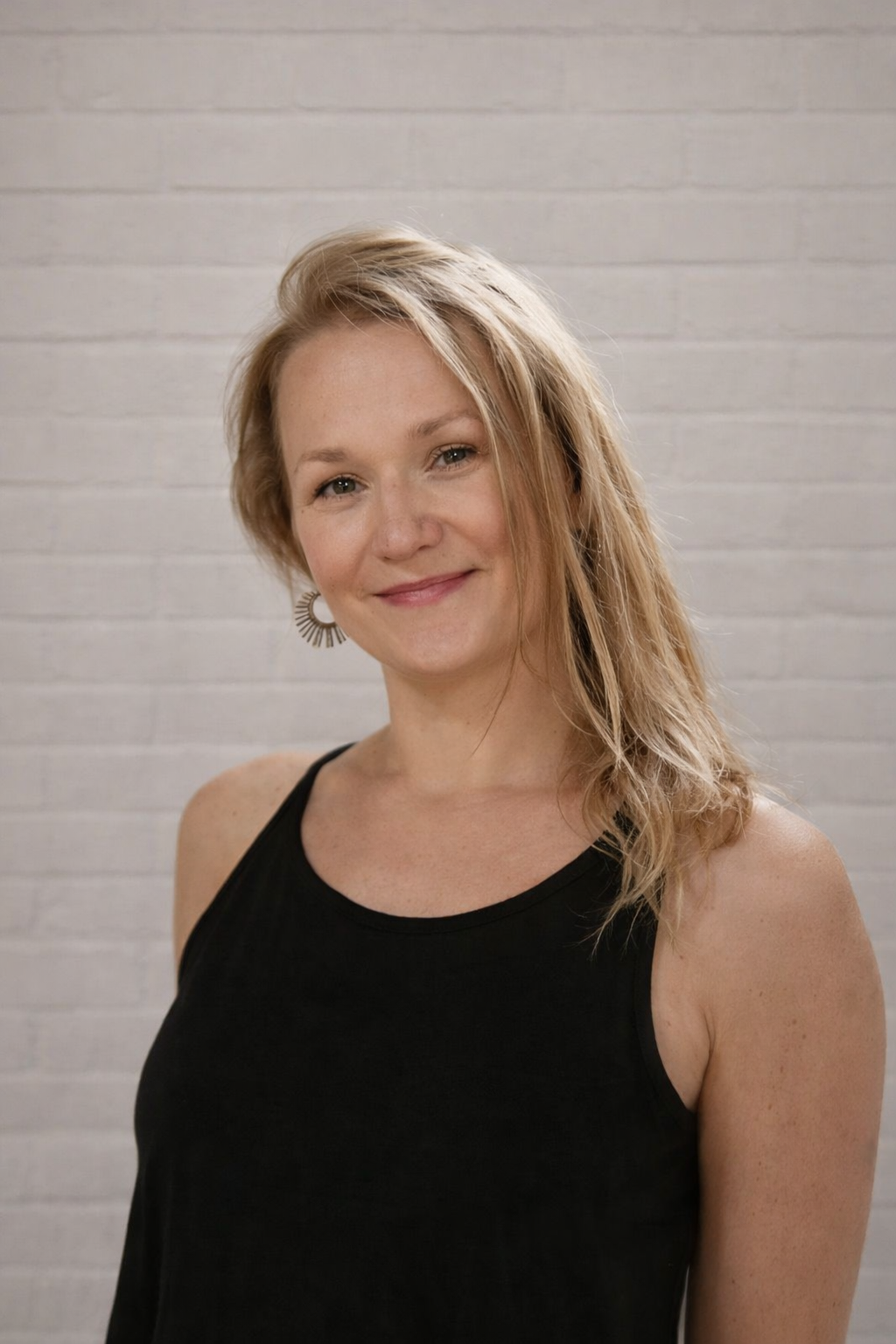 A woman with shoulder-length blonde hair wearing a black sleeveless top and earrings, smiling in front of a light brick wall.