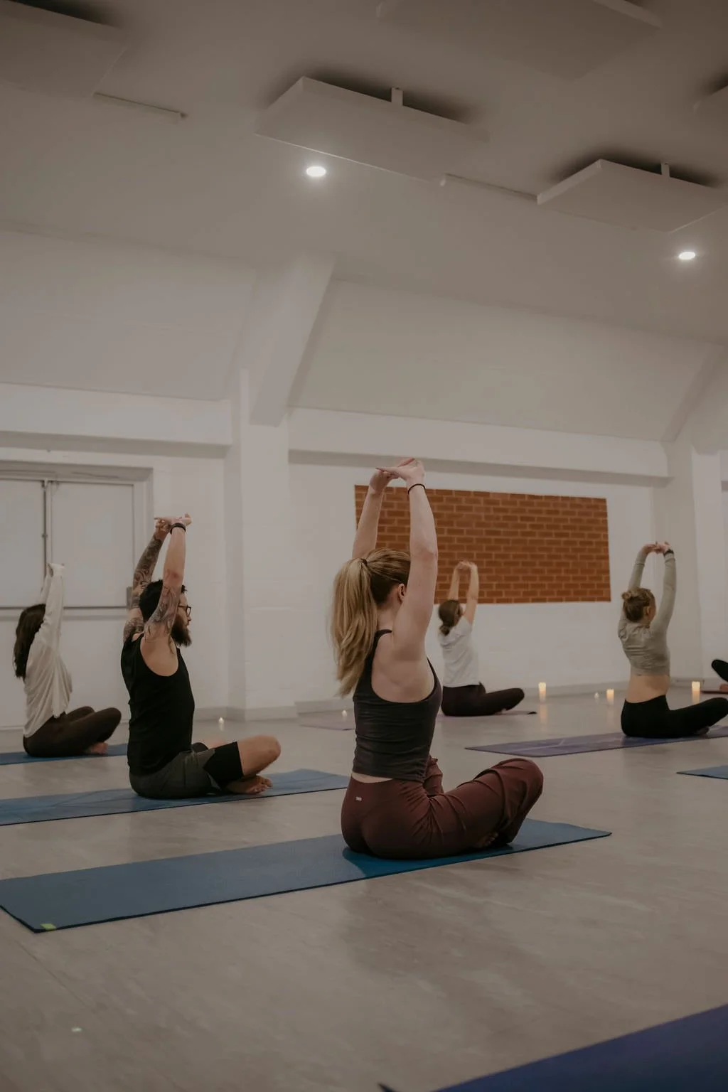 Group of people practicing yoga in a studio, sitting cross-legged with arms raised overhead.