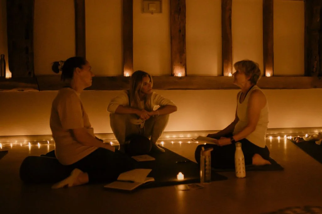 Three women sitting cross-legged on yoga mats in a dimly lit room with string lights, engaging in a conversation or meditation session.