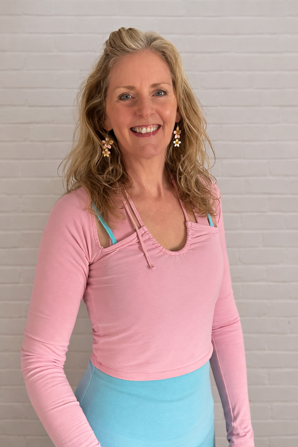 A smiling woman with blonde wavy hair, wearing pink floral earrings, a pink long-sleeve top over a blue tank top, standing in front of a white brick wall.