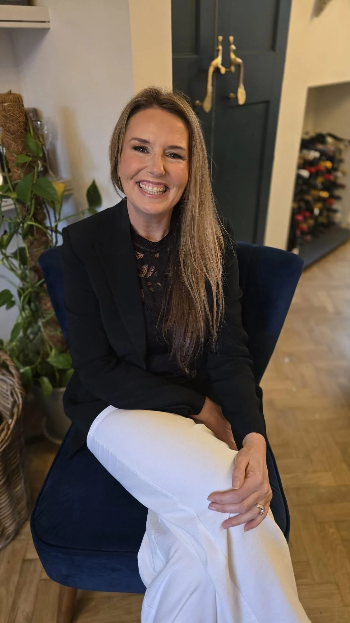 A smiling woman with long light brown hair sitting indoors. She is wearing a black blazer and light-colored pants, with a white blanket on her lap. The background shows a dark blue door with vintage brass fixtures, a wine rack, and some green plants.